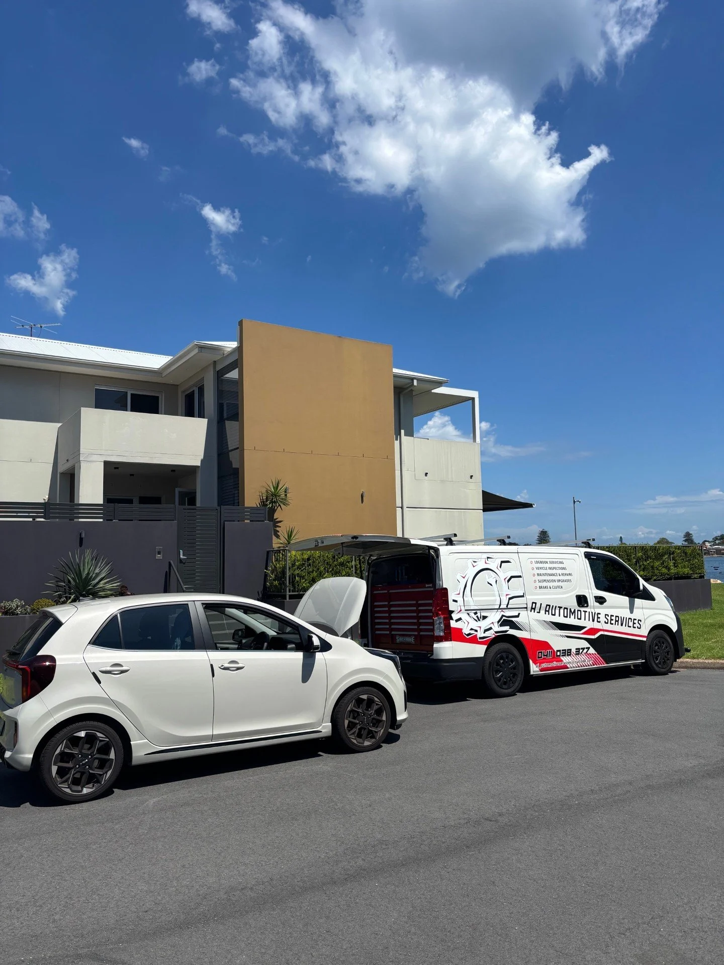 A white hatchback car and a white van with red and black branding labeled 'AJ Automotive Services' parked on a residential street with modern white apartment buildings, green plants, and a blue sky with clouds in the background.