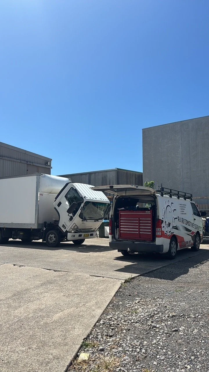 A white delivery truck with a cab tilted forward, and a service van with an open rear door showing tools inside, parked on an asphalt lot near industrial buildings.