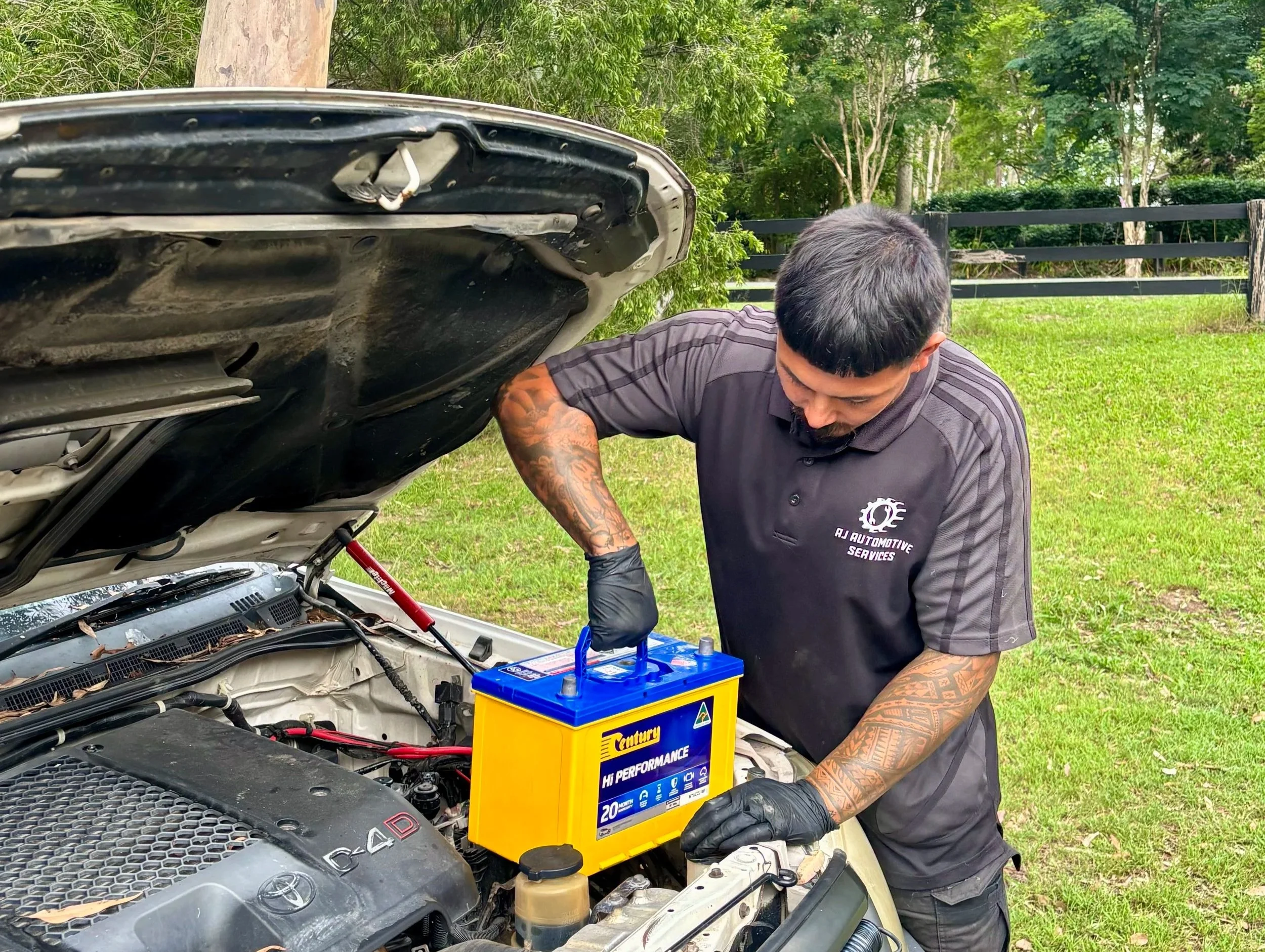 An automotive technician works on a car engine outdoors, handling a yellow battery labeled 'Century Hi Performance' with a blue handle, while wearing black gloves and a black polo shirt with an 'RJ Automotive Services' logo.