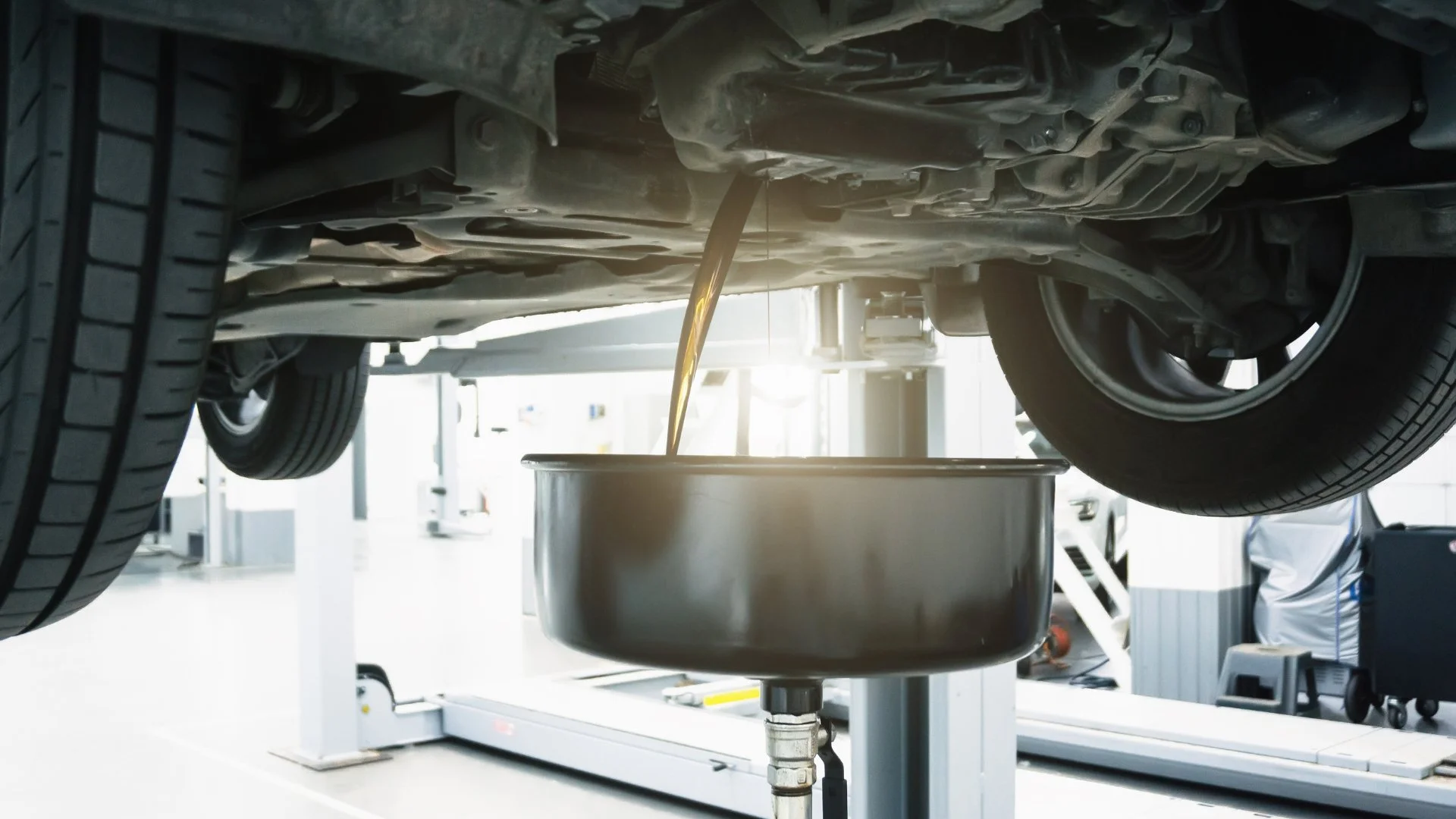 Underneath view of a car with oil being drained into a container during maintenance at an auto repair shop.