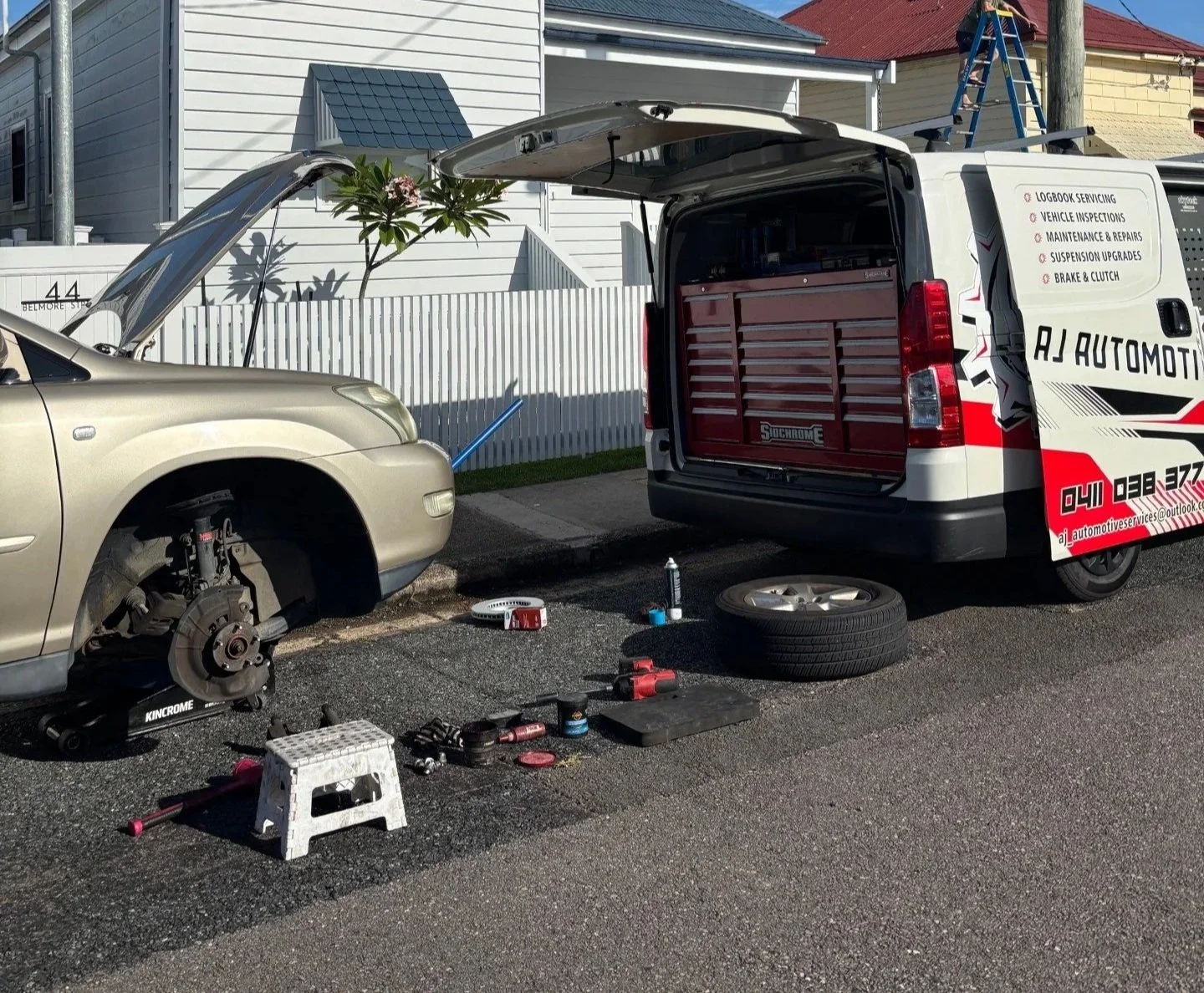 Auto repair scene with a car elevated on a jack with its front wheel removed, tools and supplies on the ground, and an open service van with equipment inside.