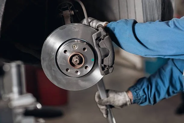 A vehicle mechanic performs maintenance on a car's brake disc and caliper, wearing a blue work uniform and gloves.