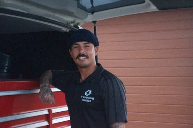 A man with a mustache and tattoos wearing a black beanie and mechanic uniform smiling, standing next to a red tool chest inside a garage.