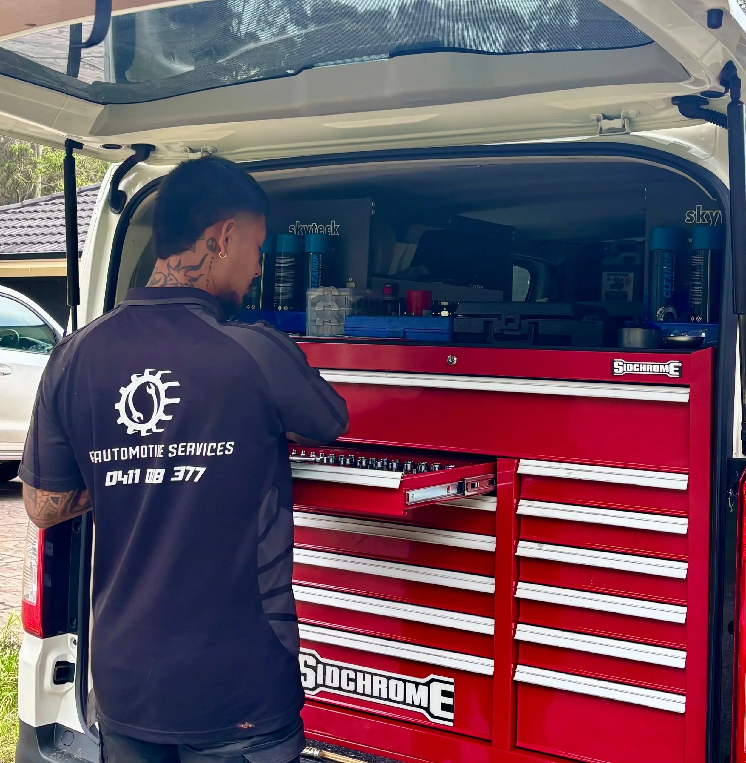 A mechanic with tattoos wearing a black uniform standing at an open mobile tool chest inside a van, working with tools on a cloudy day.