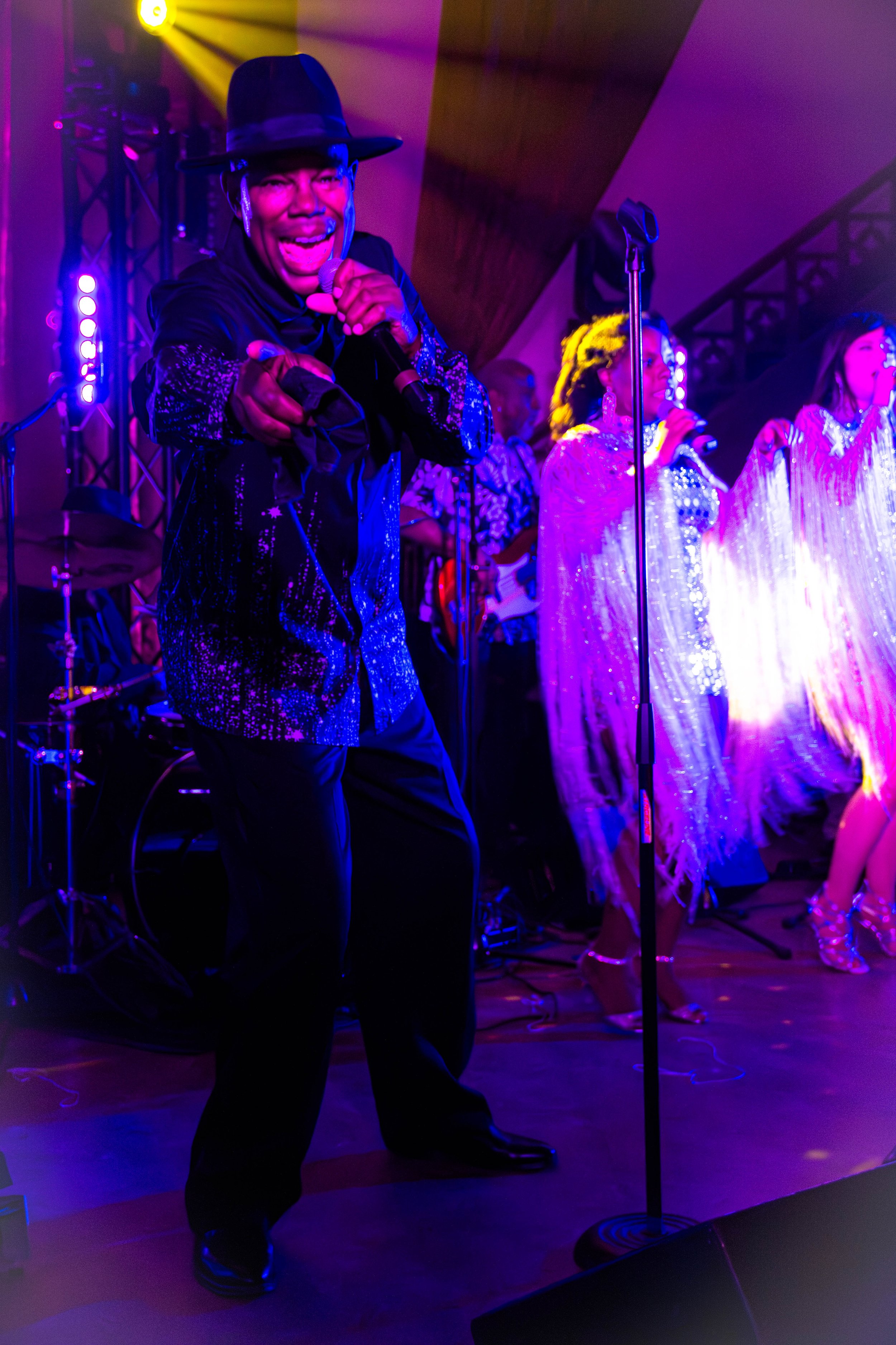 Un chanteur portant un chapeau noir et des vêtements brillants lors d'une performance musicale, avec un groupe de chanteurs et musiciens en arrière-plan, sous un éclairage violet et jaune.