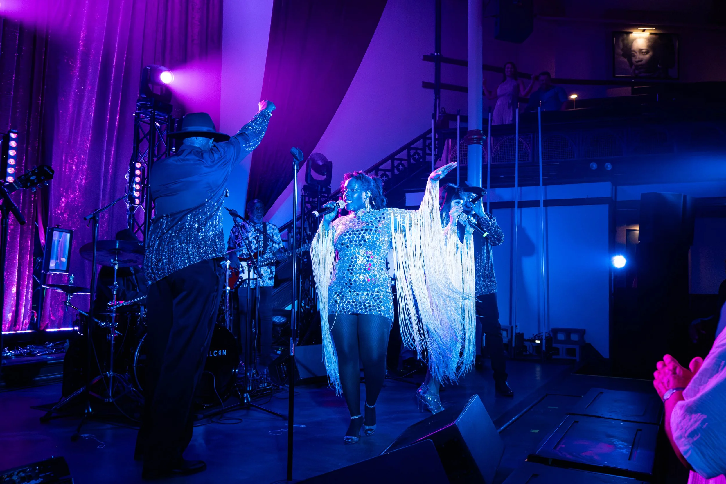 Groupe de musiciens et chanteurs en scène avec éclairage violet, une chanteuse en costume brillant avec des fringues à longues franges au centre