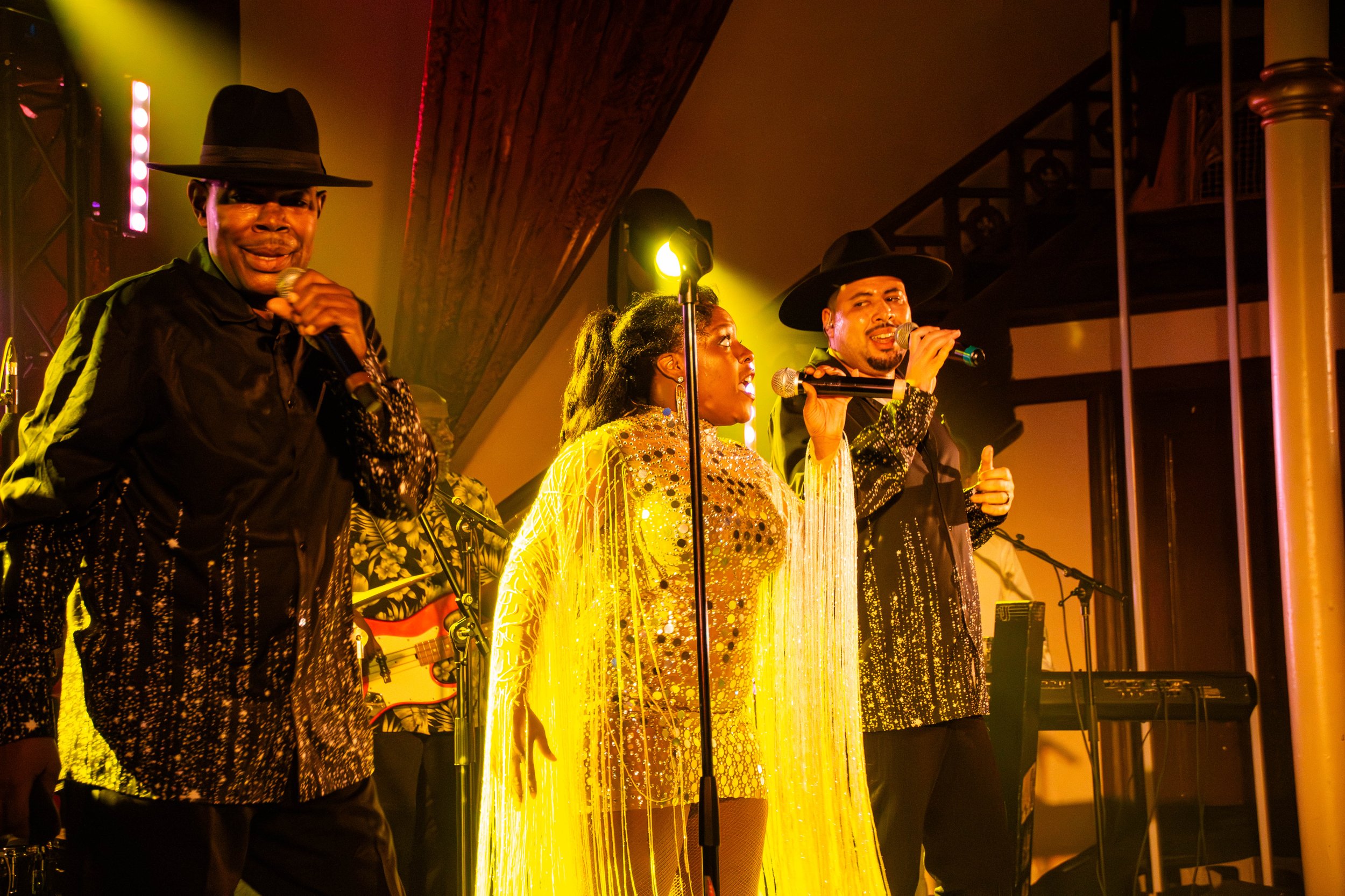 Groupe de musiciens sur scène sous éclairage jaune et orange, portants des vêtements élégants avec des chapeaux noirs.