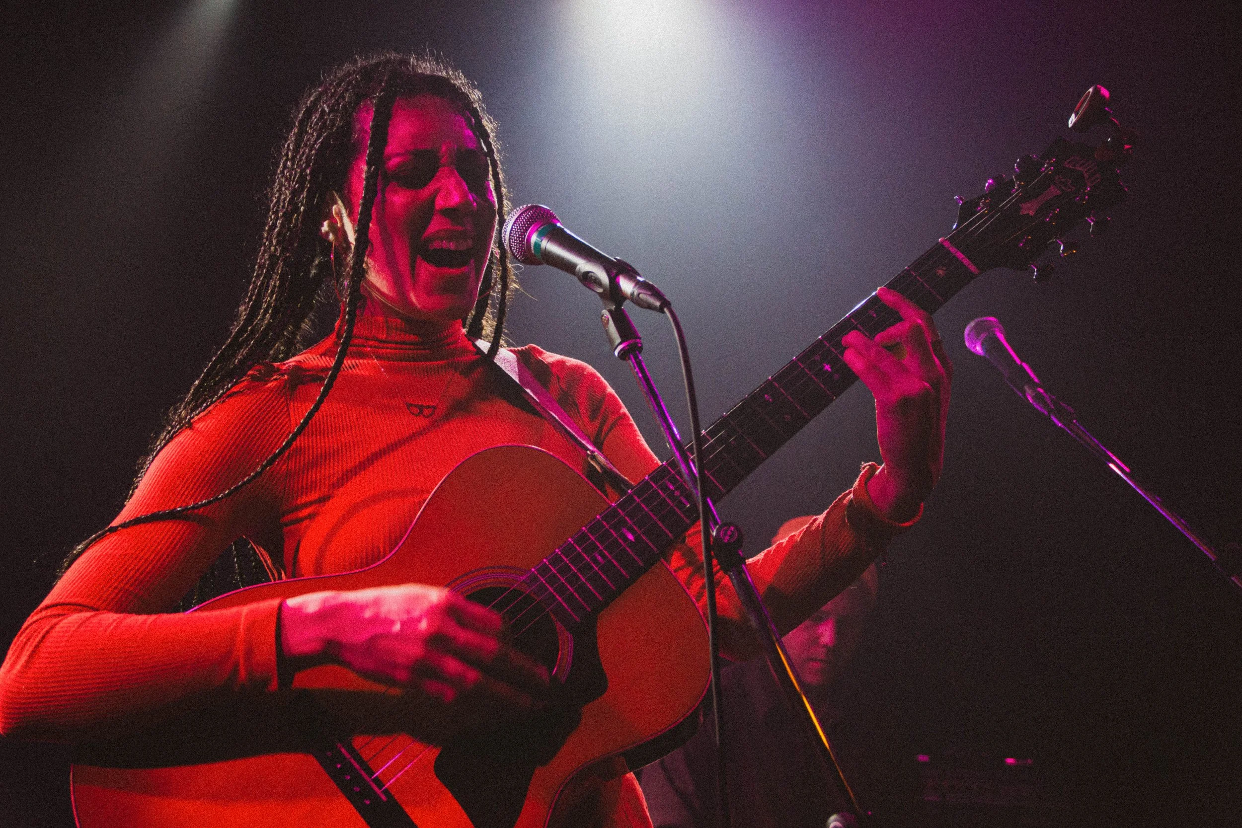 Une femme avec des dreadlocks chante et joue de la guitare sur scène, éclairée par des lumières de concert