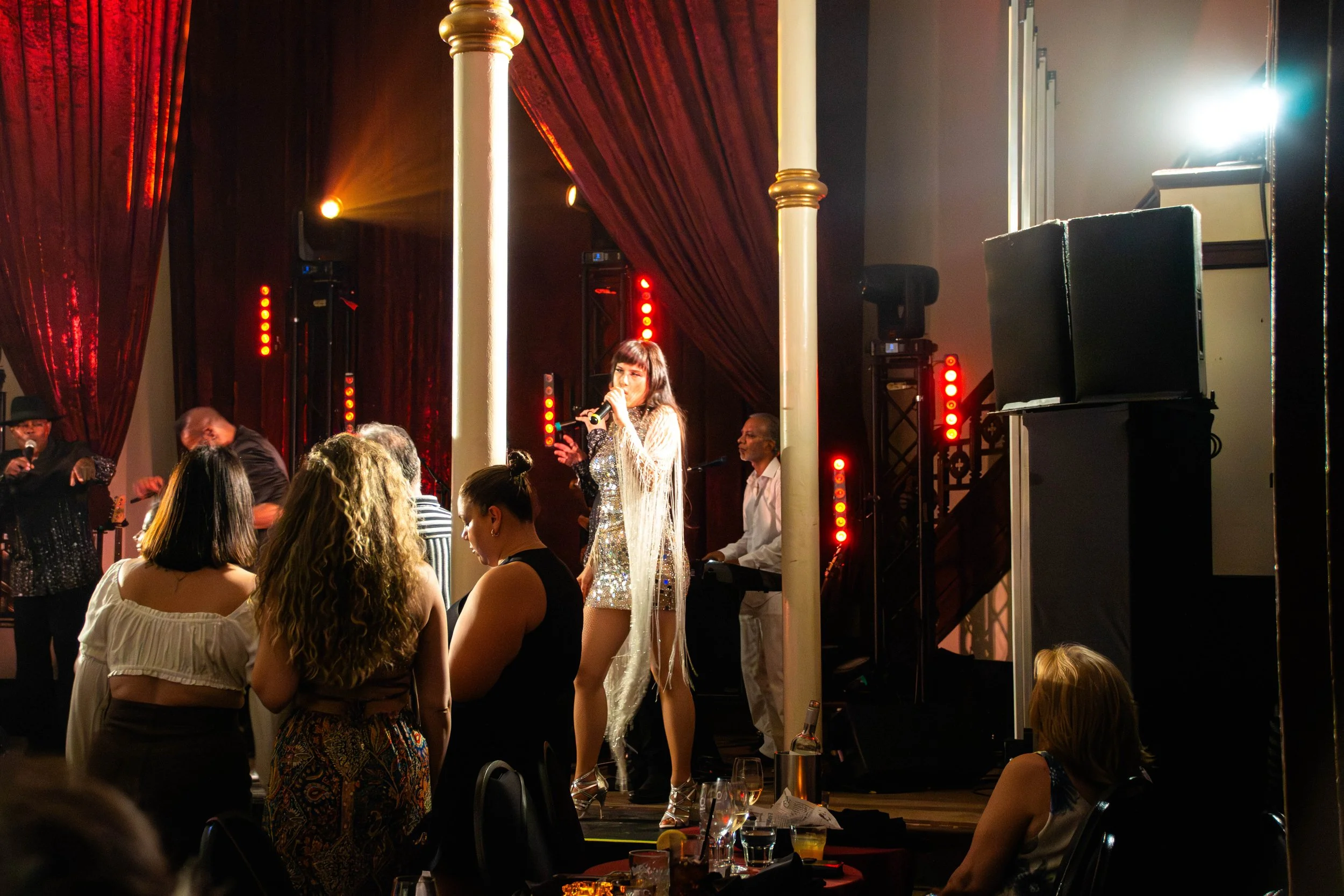 Une scène de spectacle avec une chanteuse en robe scintillante sur scène, accompagnée de musiciens, dans une salle ornée de rideaux rouges et de lumières colorées.