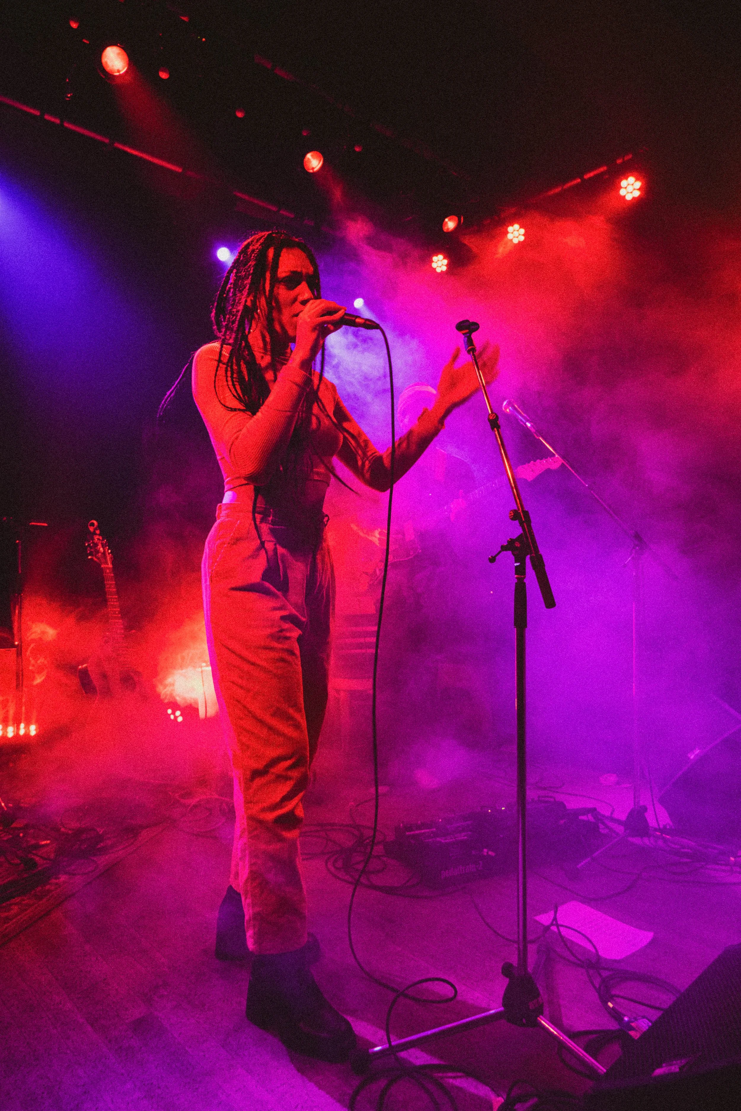 Une chanteuse aux cheveux en dreadlocks sur scène avec des lumières colorées, un microphone en main, entourée de fumée.