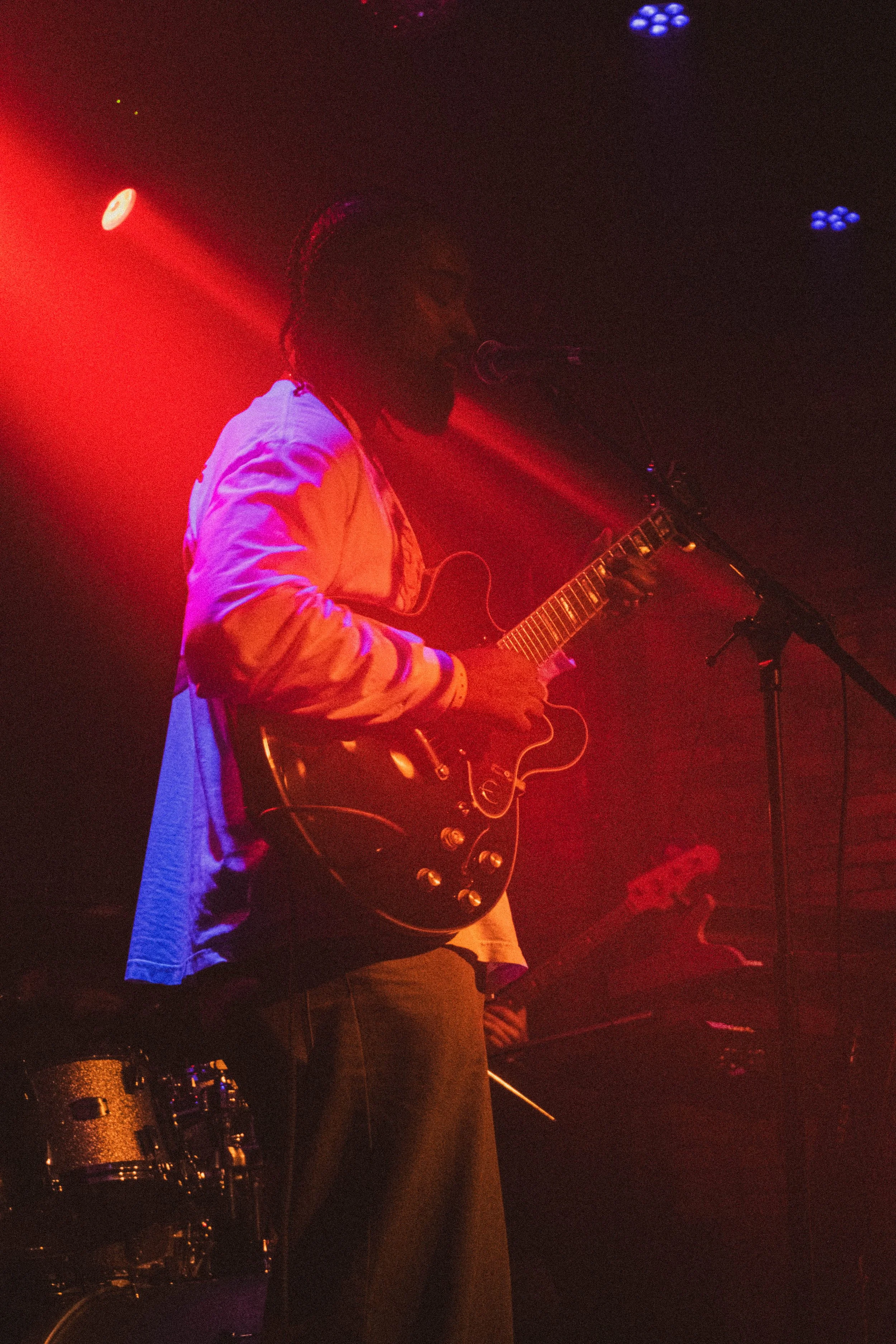 Un musicien jouant de la guitare électrique lors d'une performance sur scène, sous un éclairage rouge et violet.