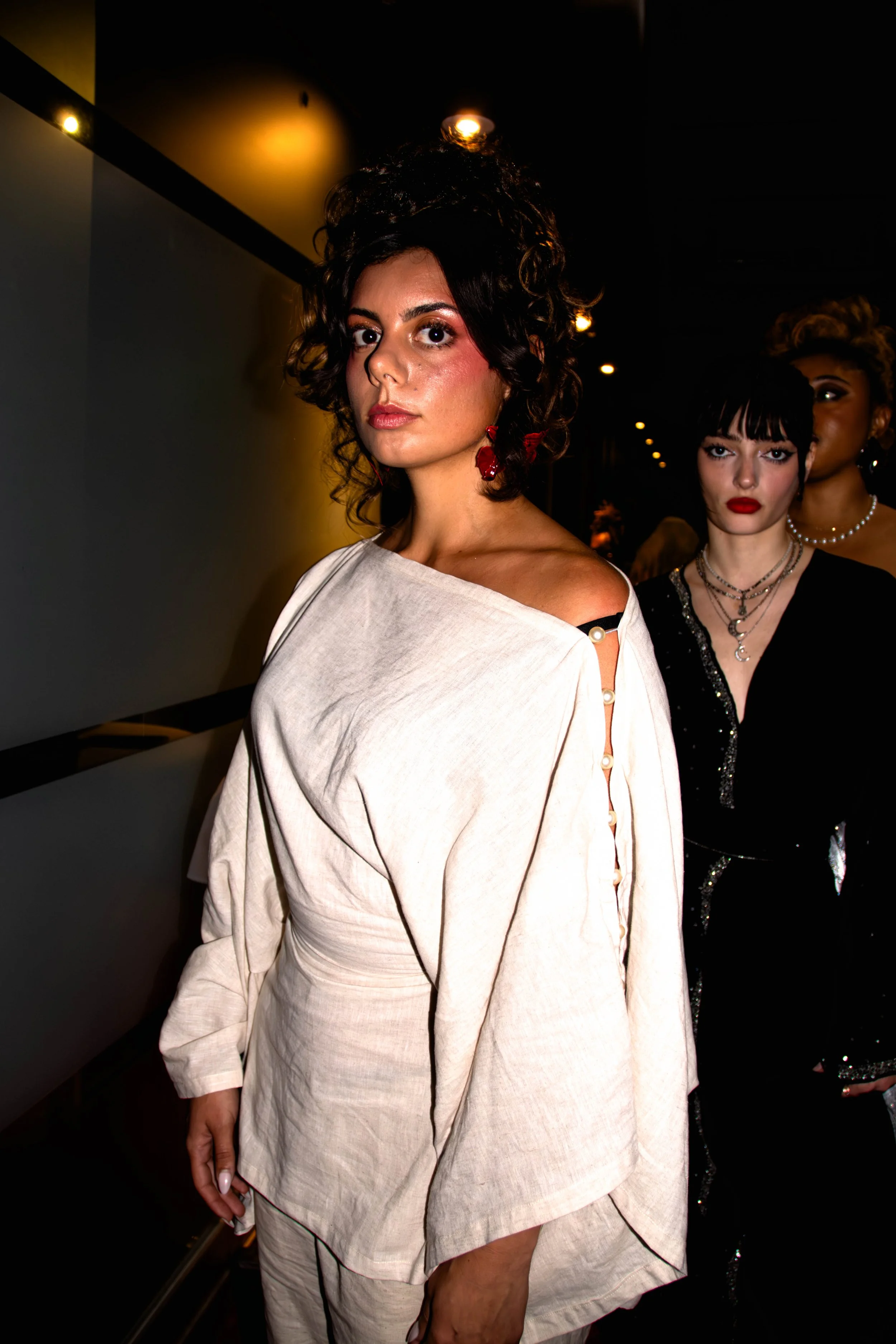 Deux femmes en vêtements élégants dans un environnement sombre, l'une porte une tenue blanche avec des détails de boutons et des boucles d'oreilles rouges, l'autre porte une robe noire avec des bijoux.