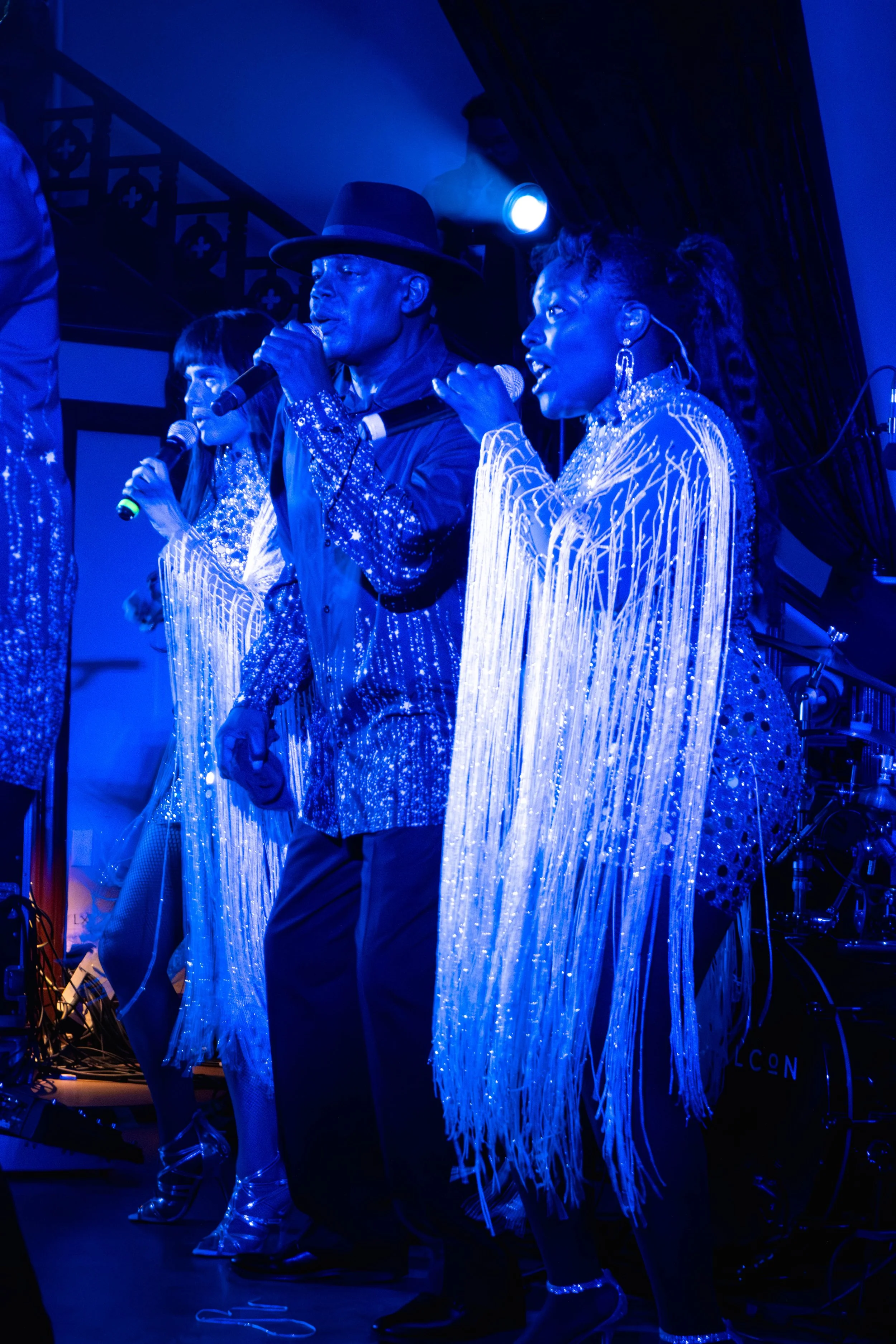 Groupe de chanteurs sur scène, vêtements brillants et lumineux sous un éclairage bleu, lors d'une performance musicale.