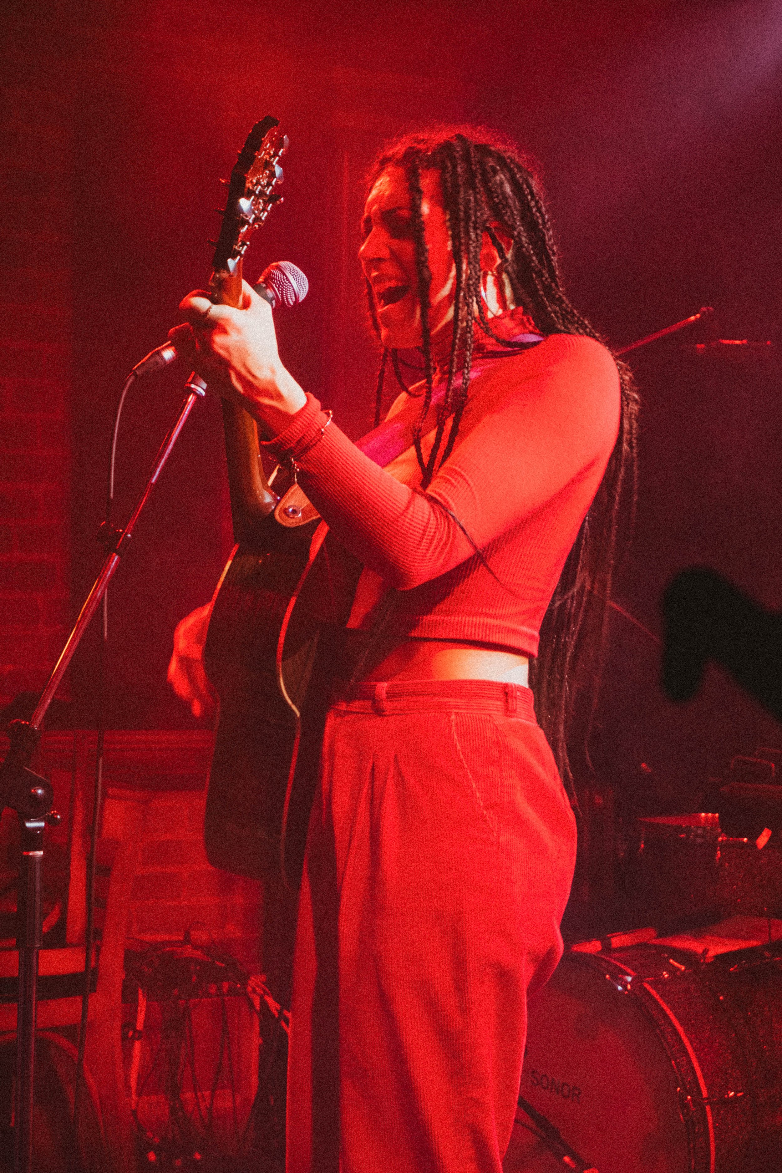 Une femme avec des dreadlocks chante en jouant de la guitare électrique lors d'une performance sur scène, nuancées de lumière rouge.