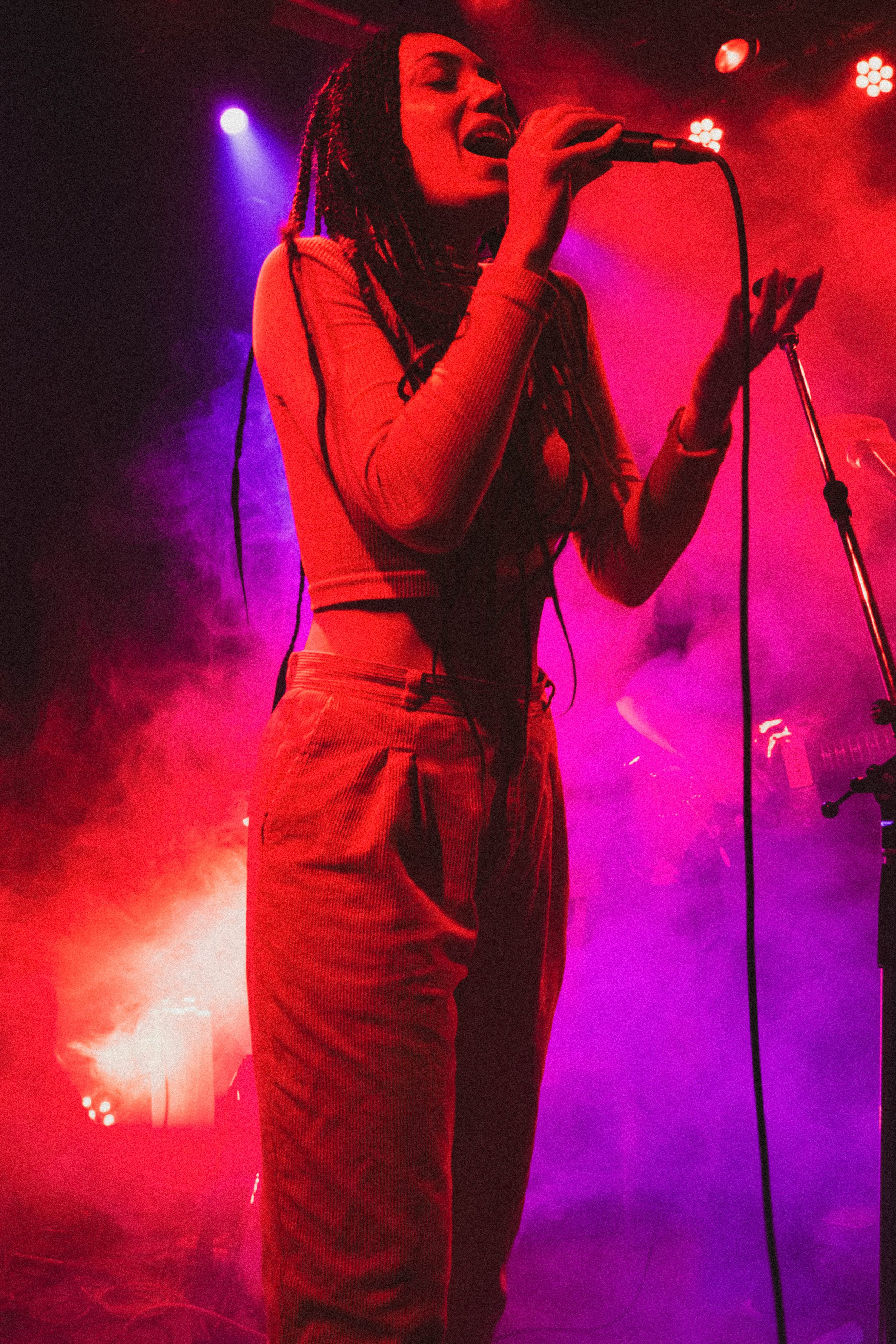 Une femme chante lors d'un concert, éclairée par des lumières violettes et rouges, avec de la fumée sur scène.