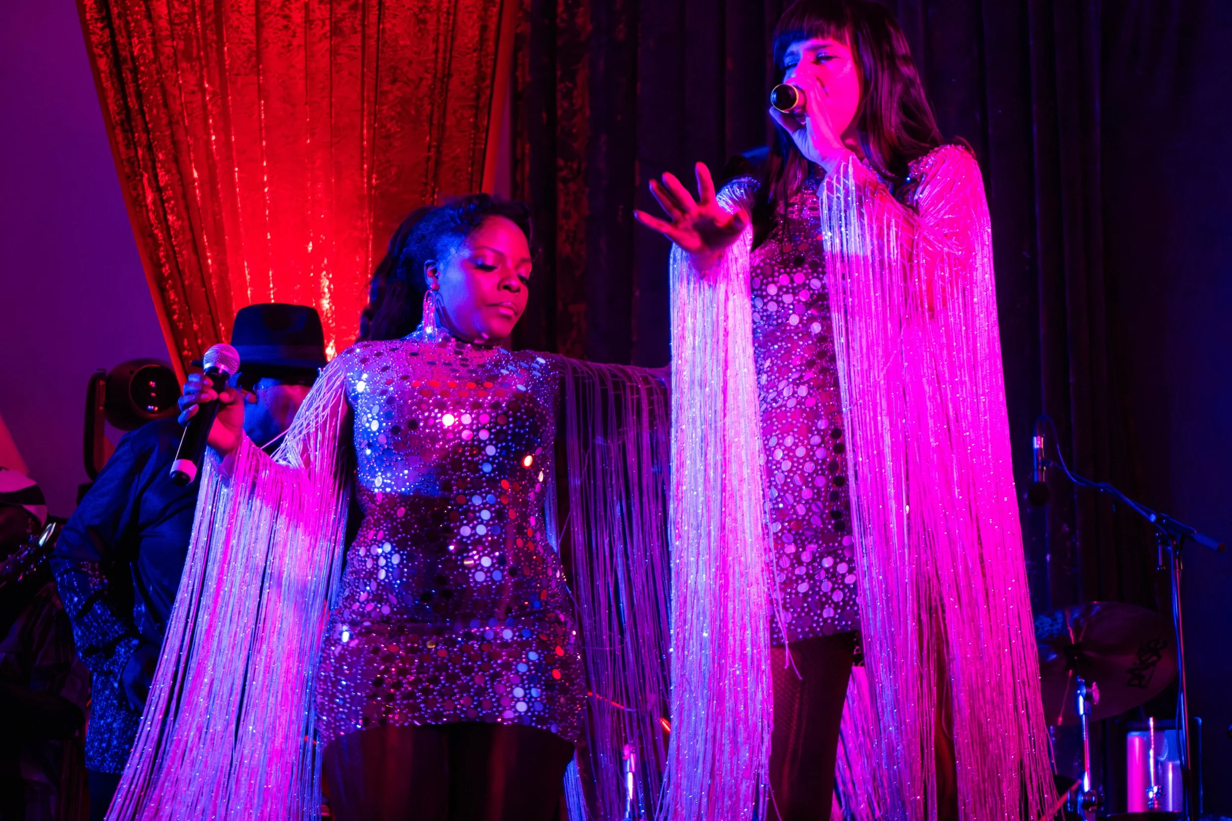 Deux femmes chantent sur scène, vêtues de robes scintillantes avec des longues rayures de strass, sous un éclairage violet et rouge, avec un fond de rideau noir et rouge.