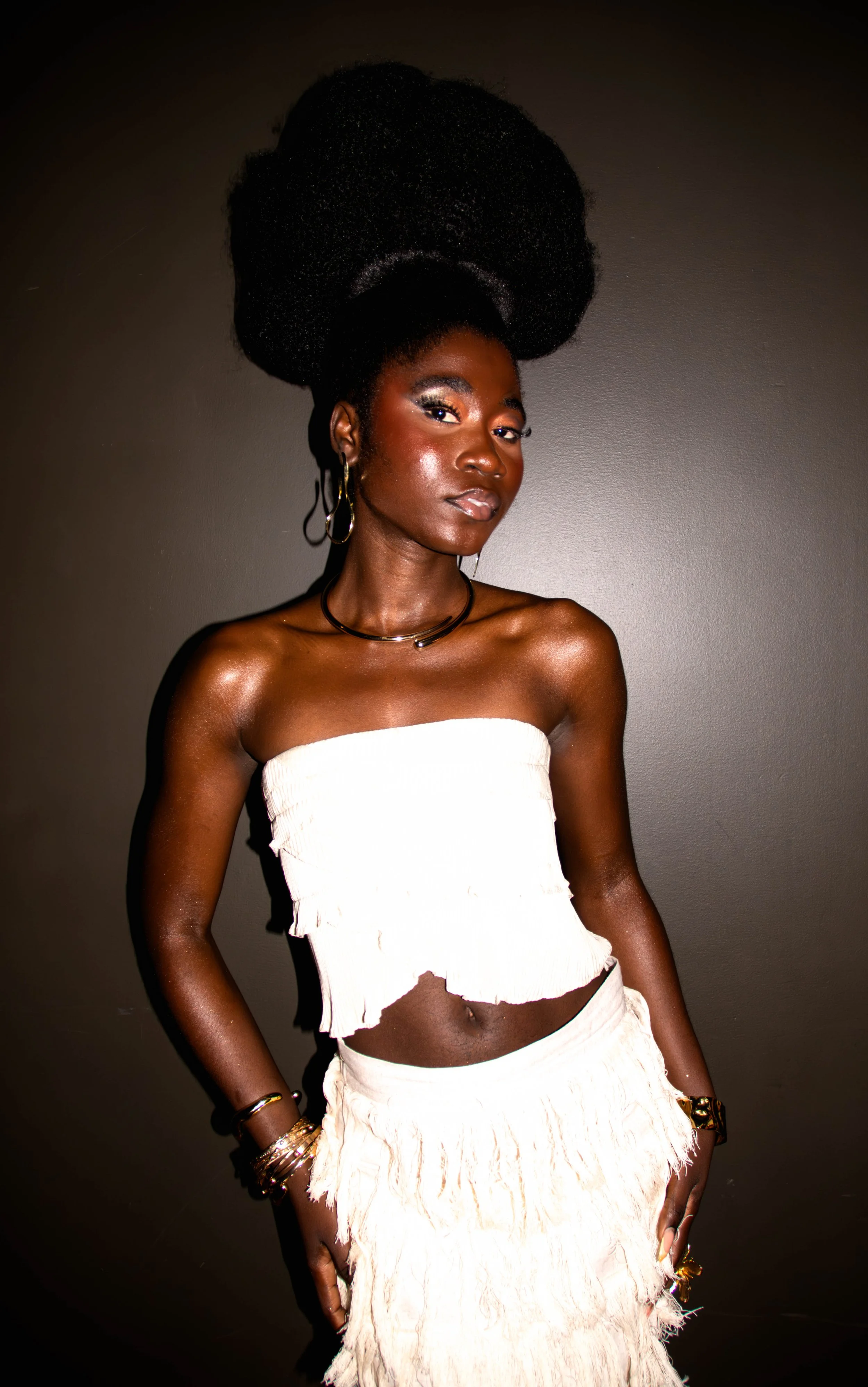 Jeune femme noire avec coiffure volumineuse, portant des vêtements blancs décontractés, accessoires dorés, armorée, regardant l'objectif devant un fond gris