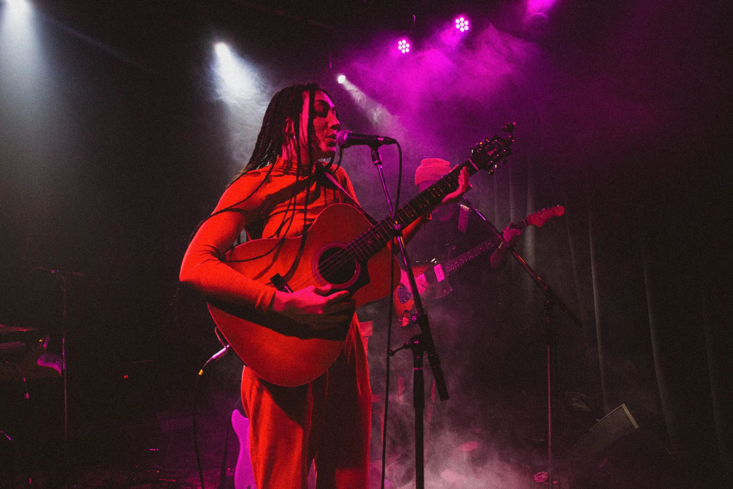 Une femme joue de la guitare acoustique lors d'un concert, entourée de lumières violettes et de fumée, avec un homme en arrière-plan portant un chapeau.