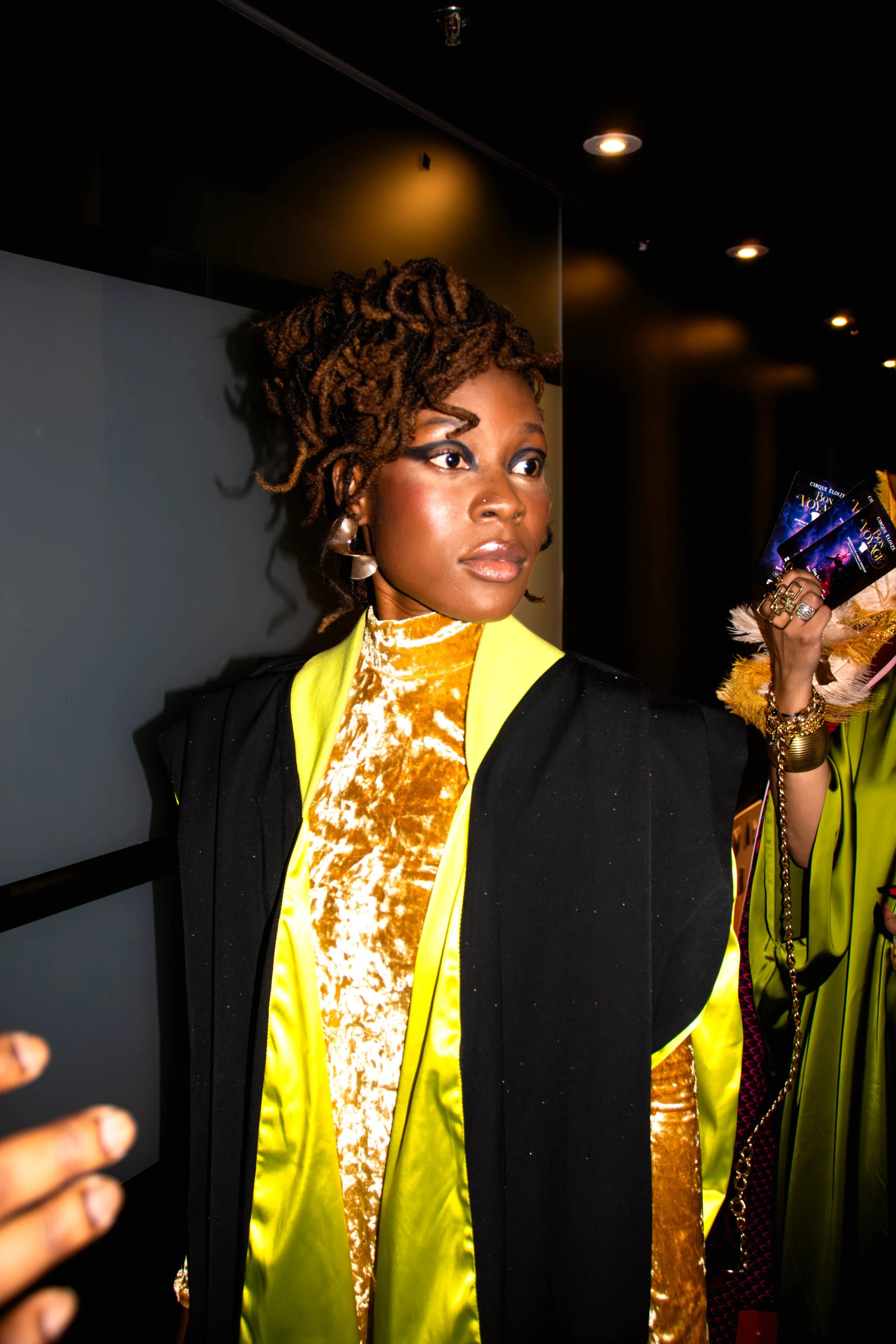 Une femme avec des dreadlocks bruns, portant une veste noire avec un intérieur jaune fluo, une robe en velours dorée et des boucles d'oreilles en forme de ruban, elle tient des billets de spectacle dans la main, dans un environnement lumineux.