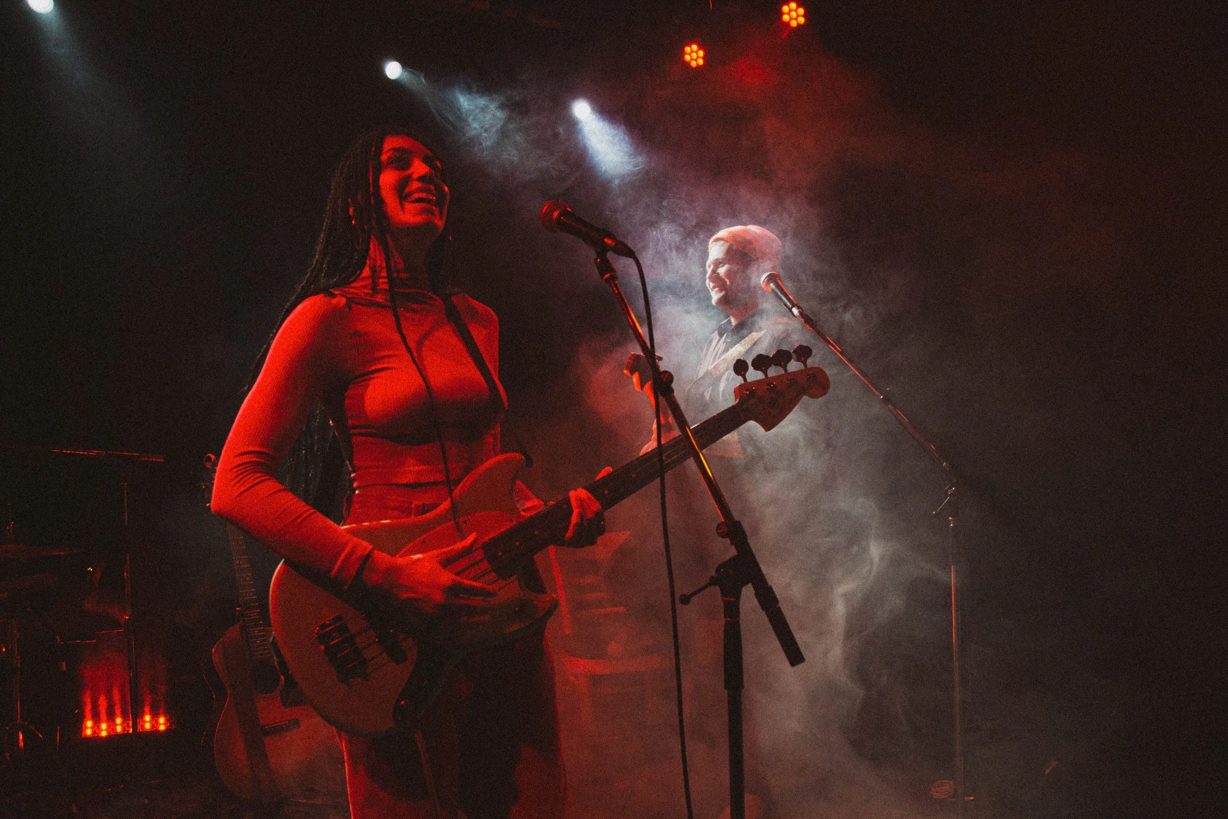 Deux musiciens jouent sur scène avec des microphones, sous des lumières rouges et blanches, entourés de fumée.