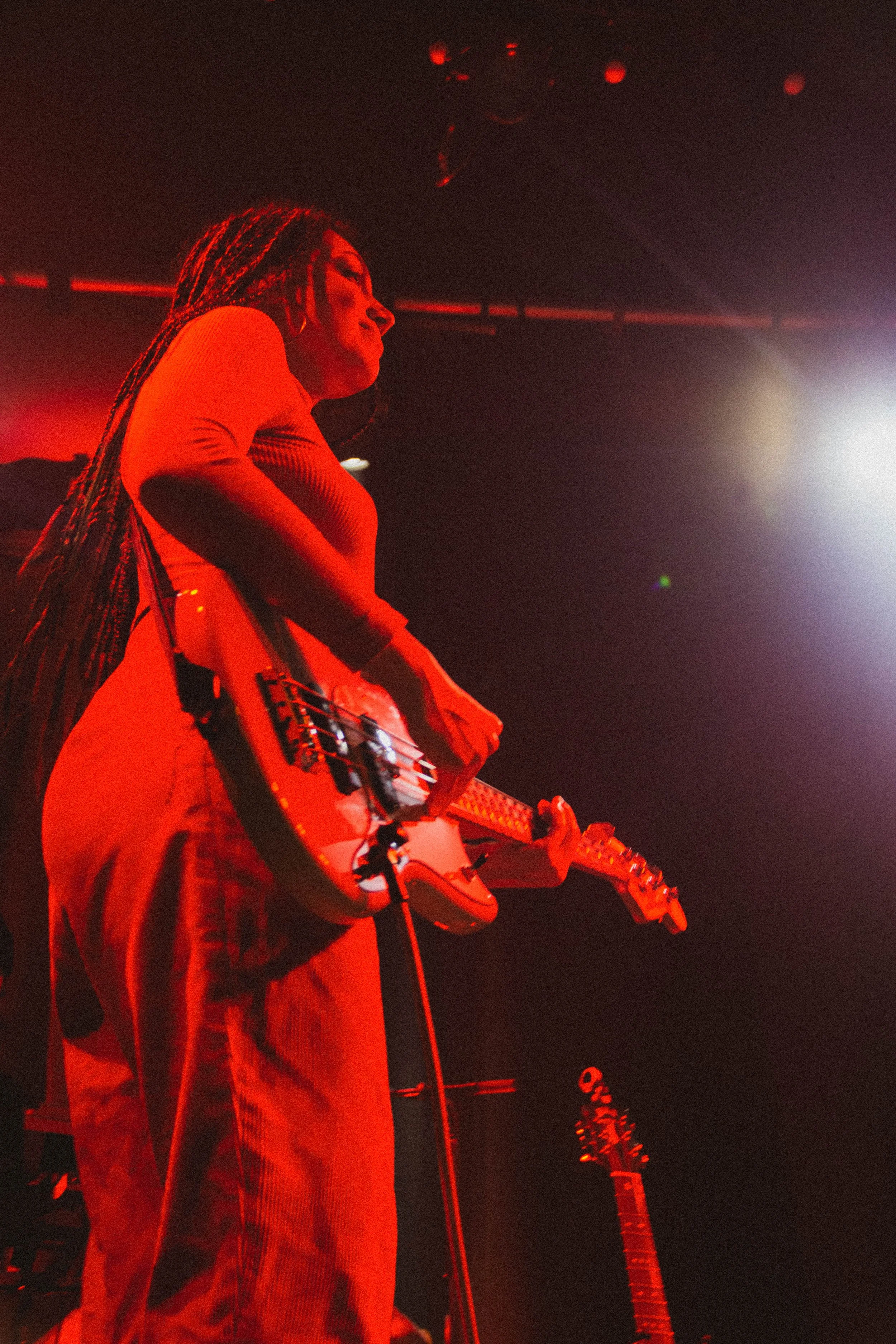 Une femme joue de la guitare électrique sur scène sous un éclairage rouge et violet.