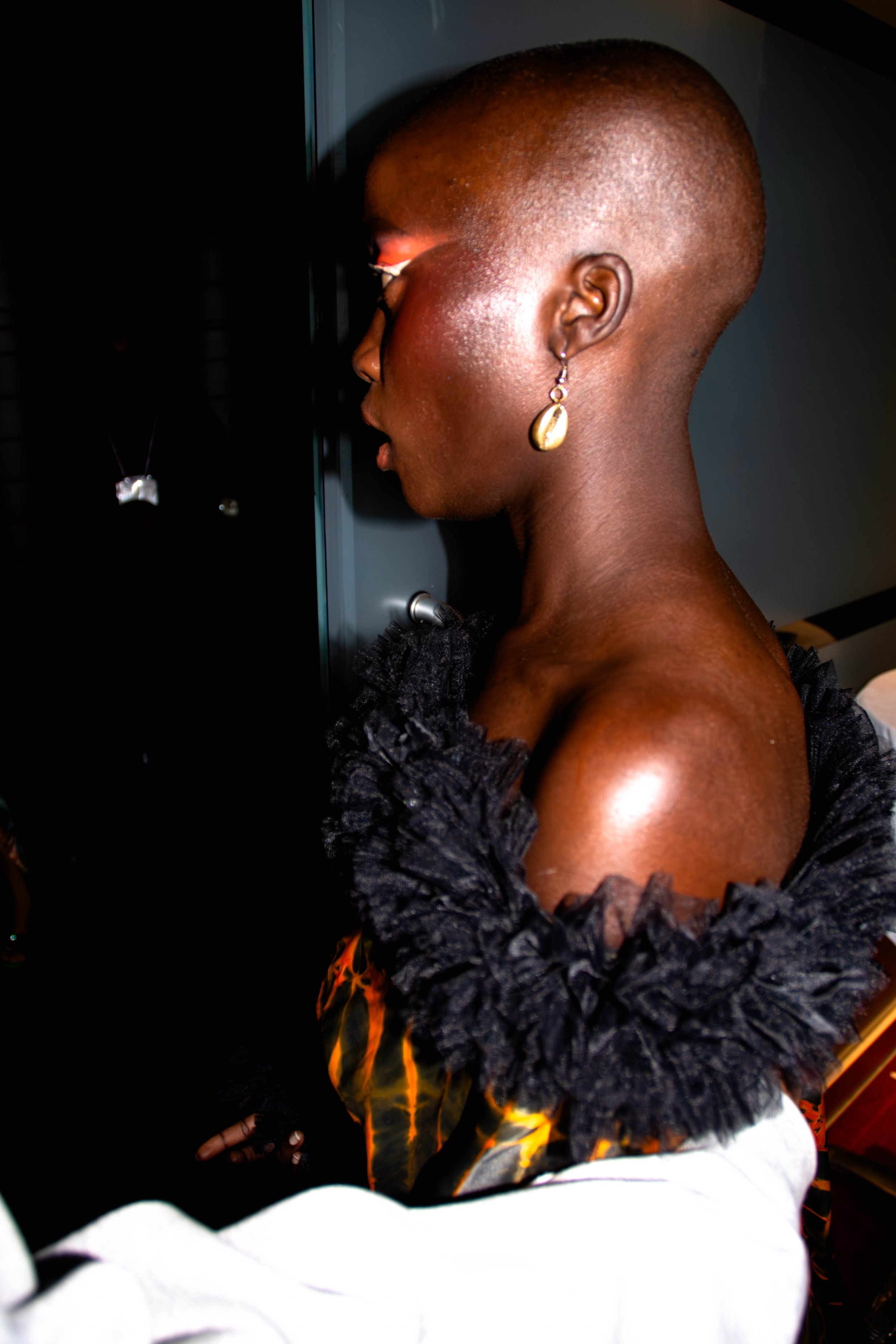 Une femme noire avec une tête rasée, portant une boucle d'oreille dorée, regardant vers un casier ou un mur avec des bijoux et vêtements suspendus.