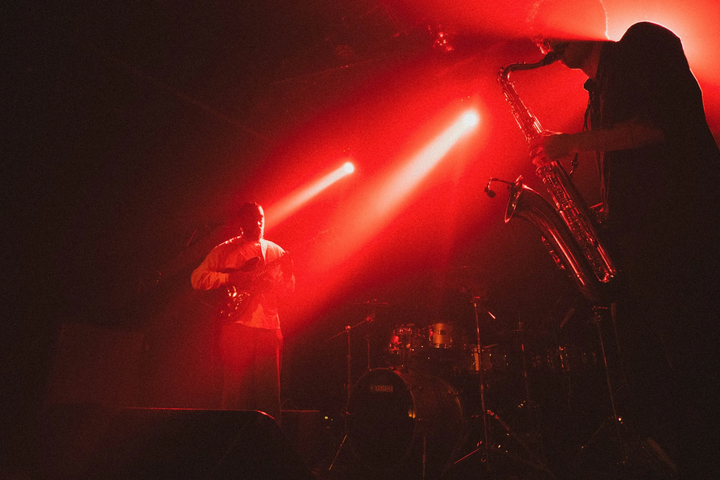 Deux musiciens jouent sur scène sous un éclairage rouge, l'un avec une guitare et l'autre avec un saxophone, avec un fond de batterie.
