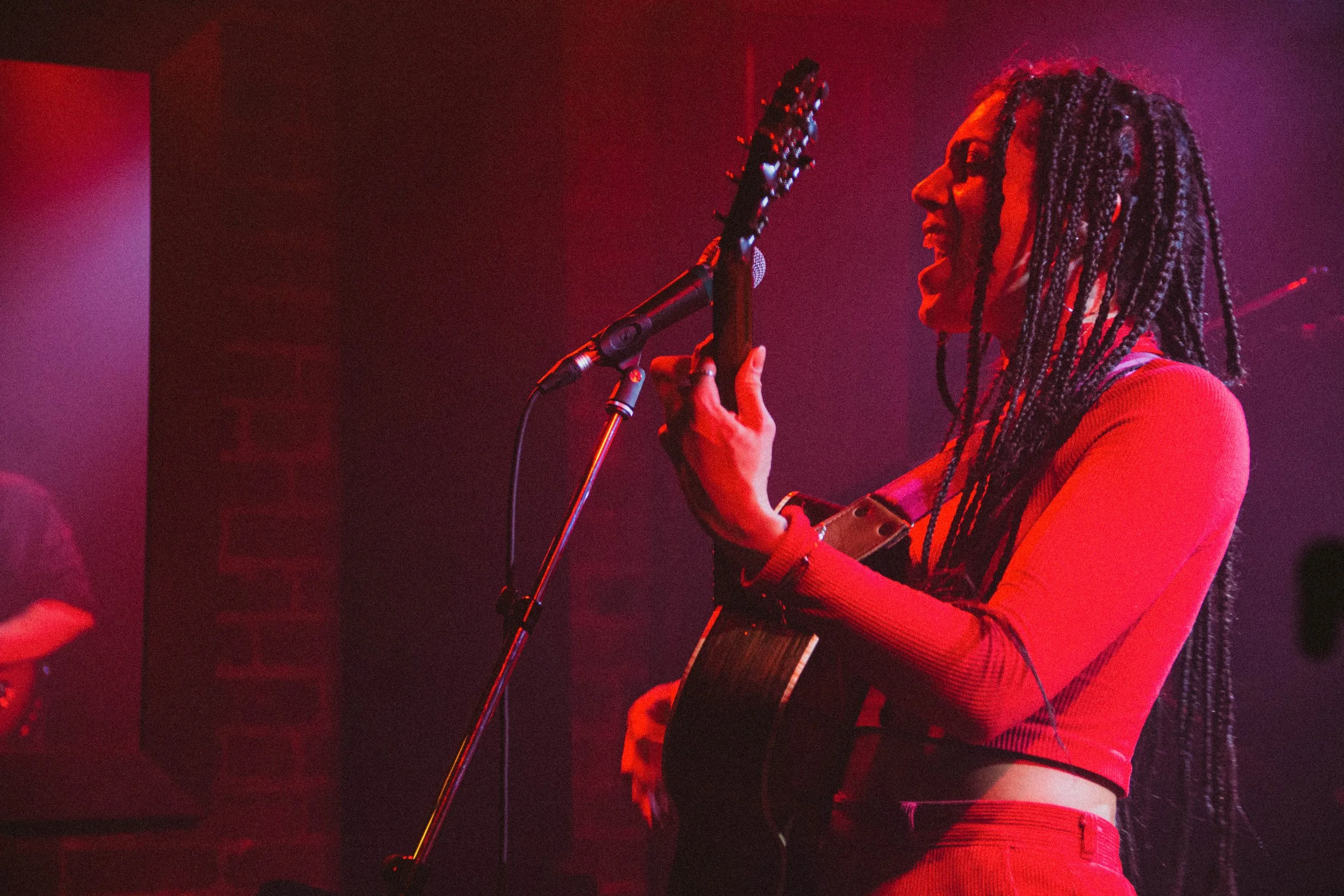 Une femme aux cheveux en dreadlocks joue de la guitare et chante dans une scène sombre avec un éclairage rouge.