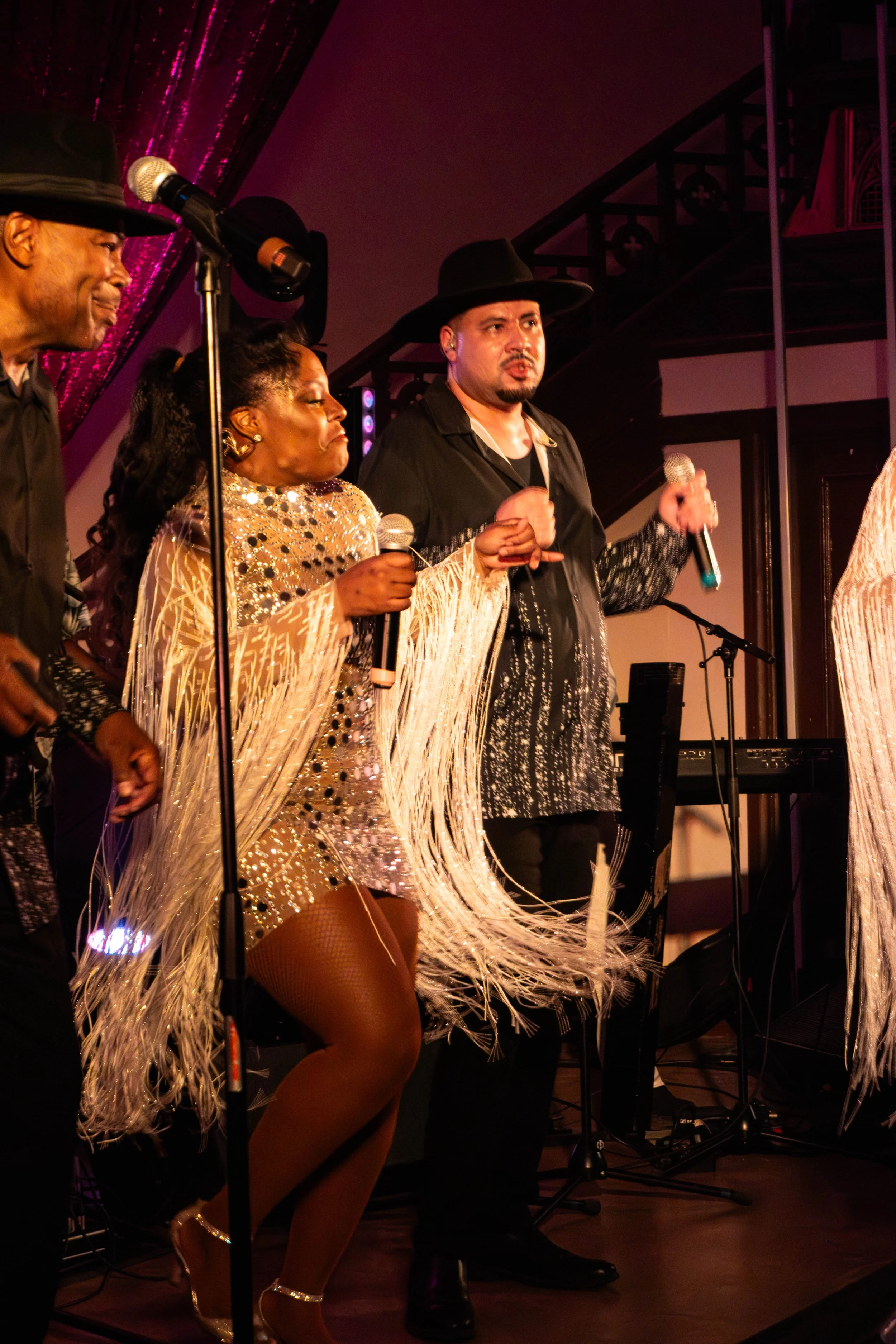 Groupe de musiciens en performance sur scène, vêtus de costumes scintillants et robustes, avec microphones, sous un éclairage chaud dans un cadre intérieur.