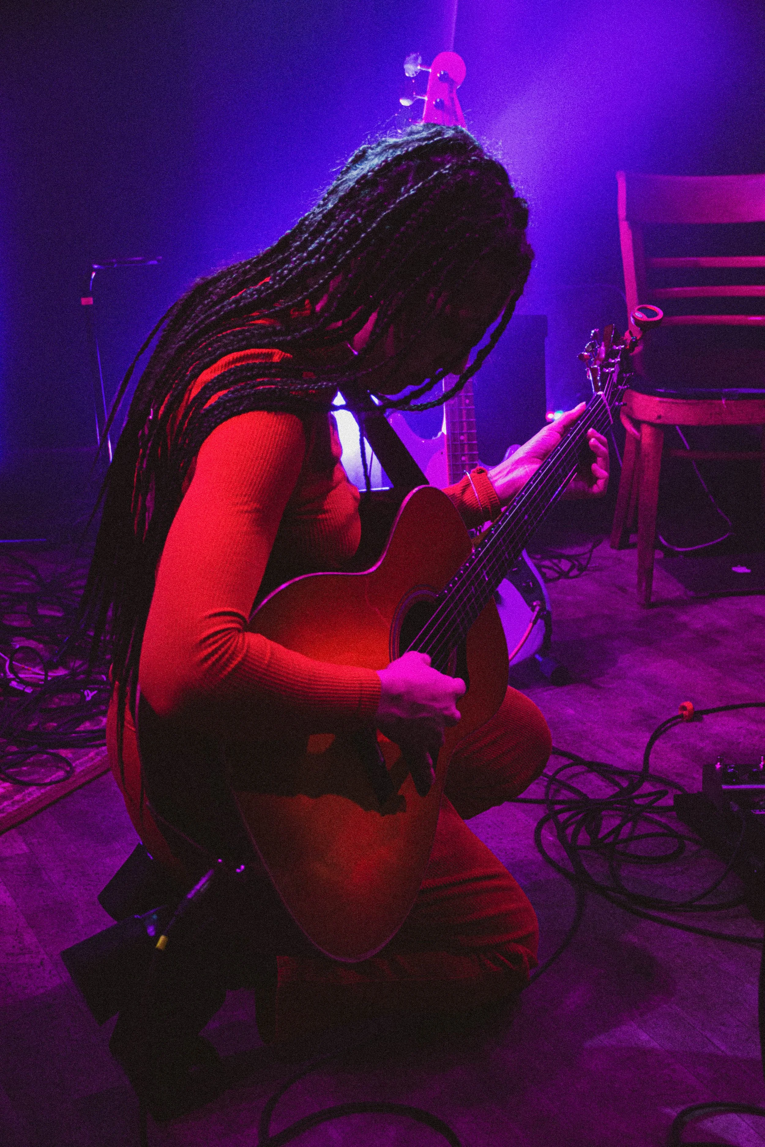 Une personne jouant de la guitare acoustique sur scène, sous un éclairage violet et rouge, avec un casque audio sur la tête.