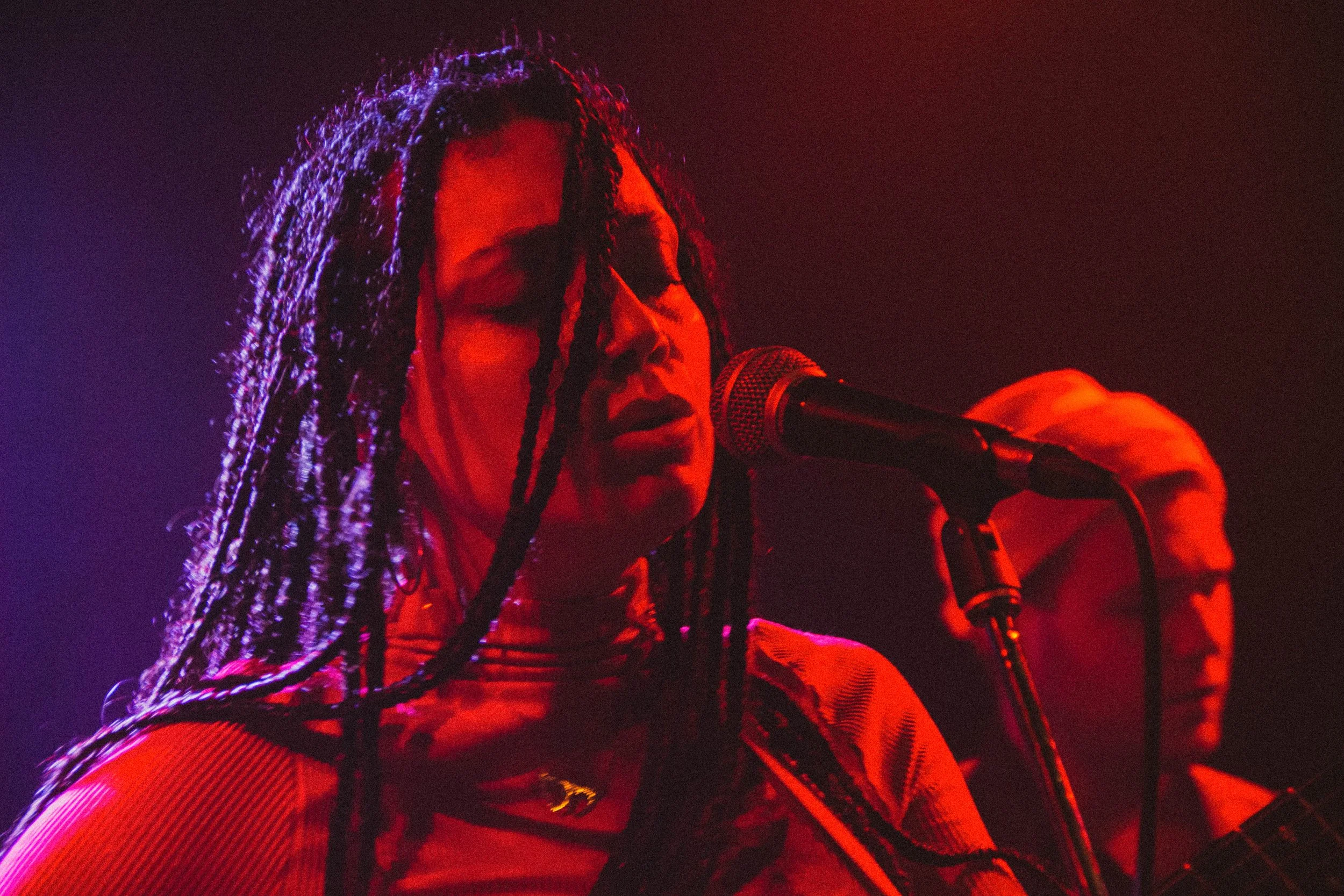 Une femme avec des cheveux en dreadlocks chante ou parle dans un microphone, avec un homme à l'arrière jouant de la guitare, sous un éclairage rouge et violet.