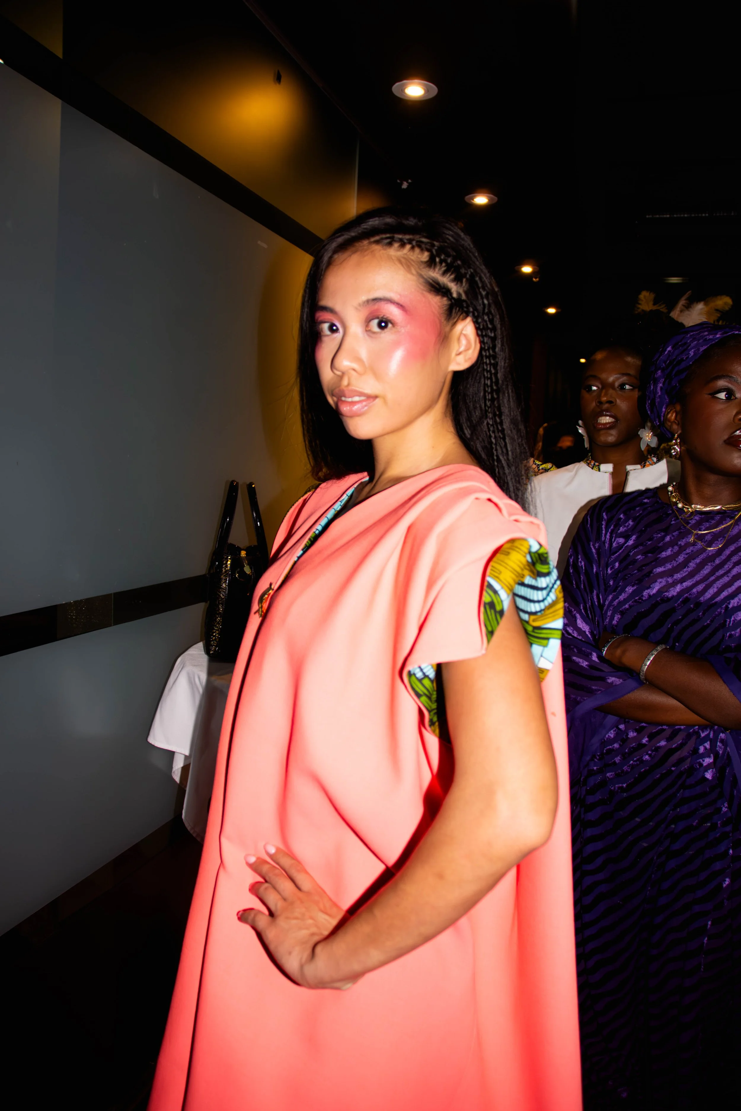 Une femme portant une robe rose avec des détails colorés sur les épaules se tient dans une pièce sombre avec d'autres personnes en arrière-plan.