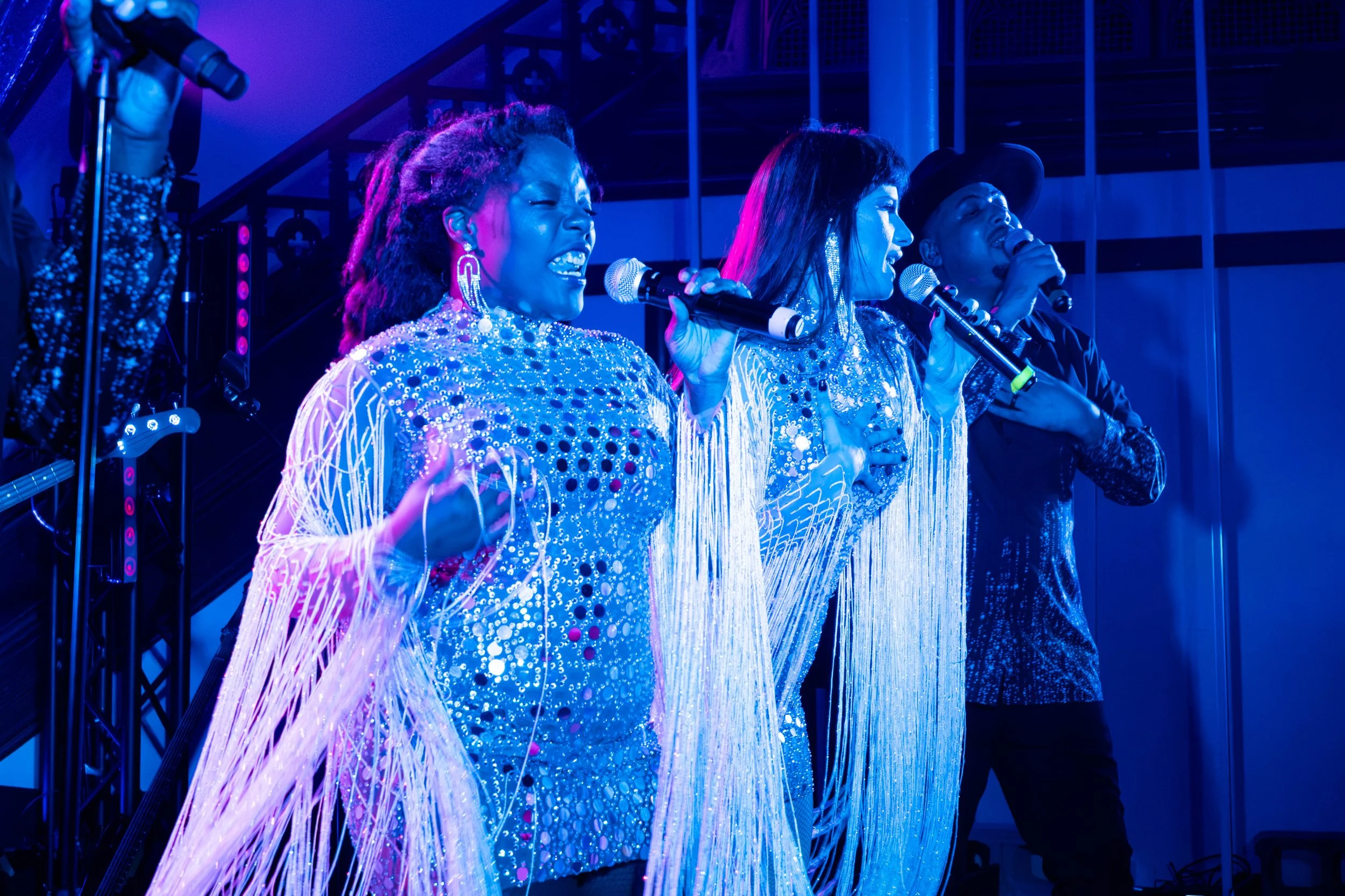Quatre chanteurs, deux femmes et deux hommes, chantent sur scène sous des lumières bleues, vêtus de costumes brillants et de vêtements à franges, tenant des microphones.