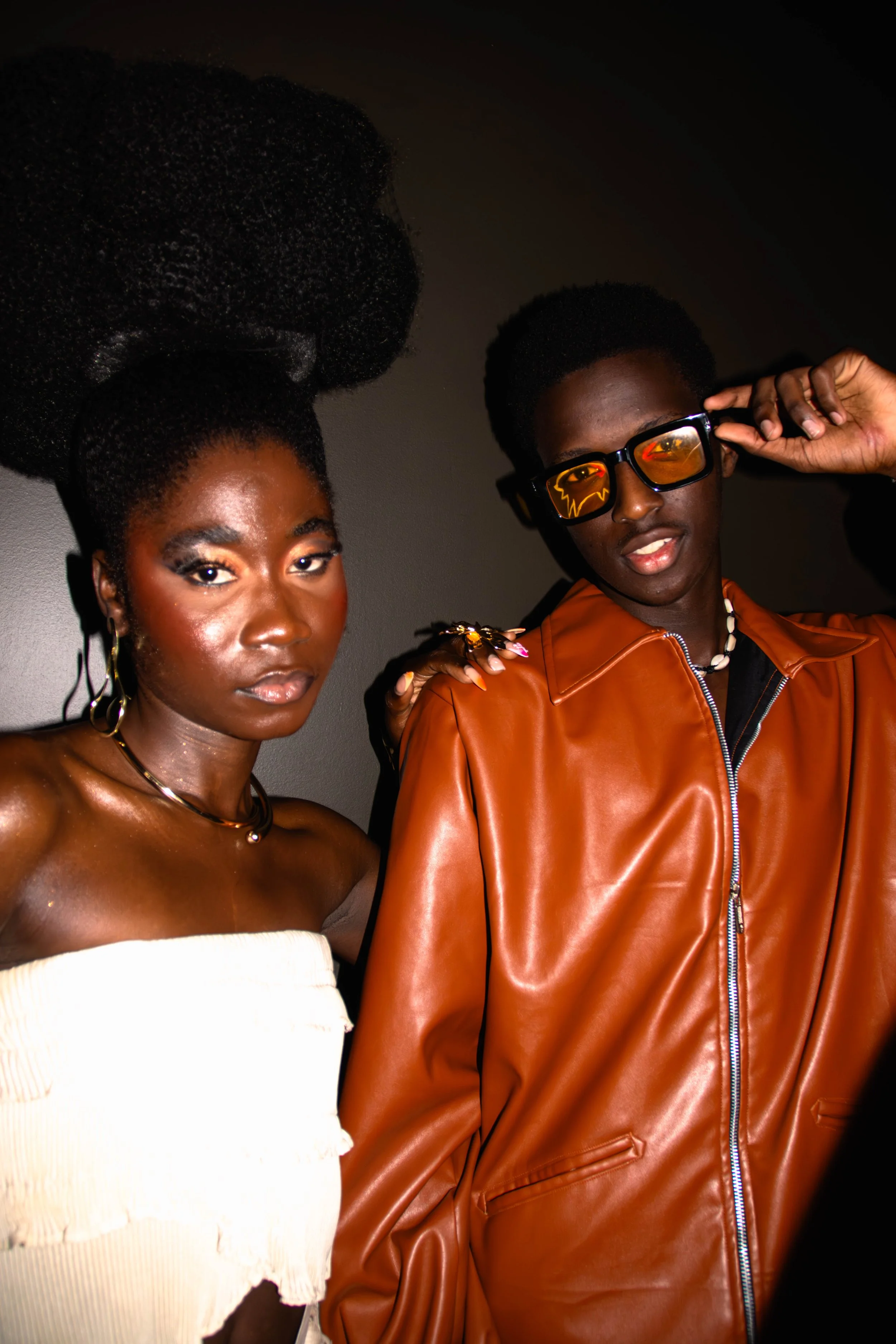 Deux jeunes noirs posent côte à côte, elle porte une robe blanche minimale et des bijoux, lui porte une veste en cuir marron clair et des lunettes de soleil.