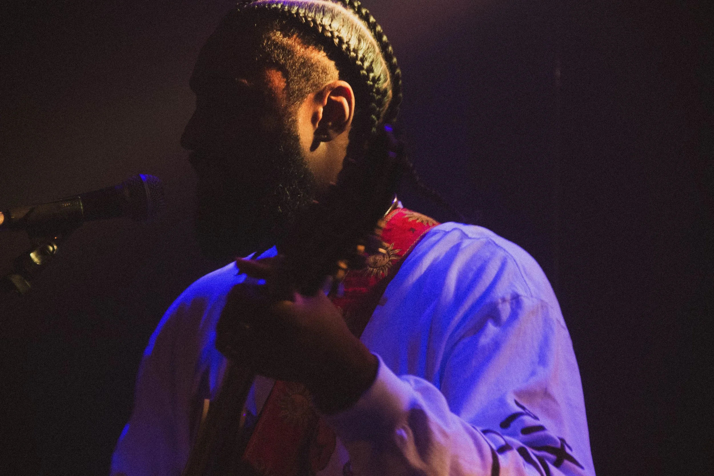 Un homme avec des tresses et une barbe, portant une robe blanche, près d'un microphone dans une ambiance sombre, probablement lors d'une performance musicale.