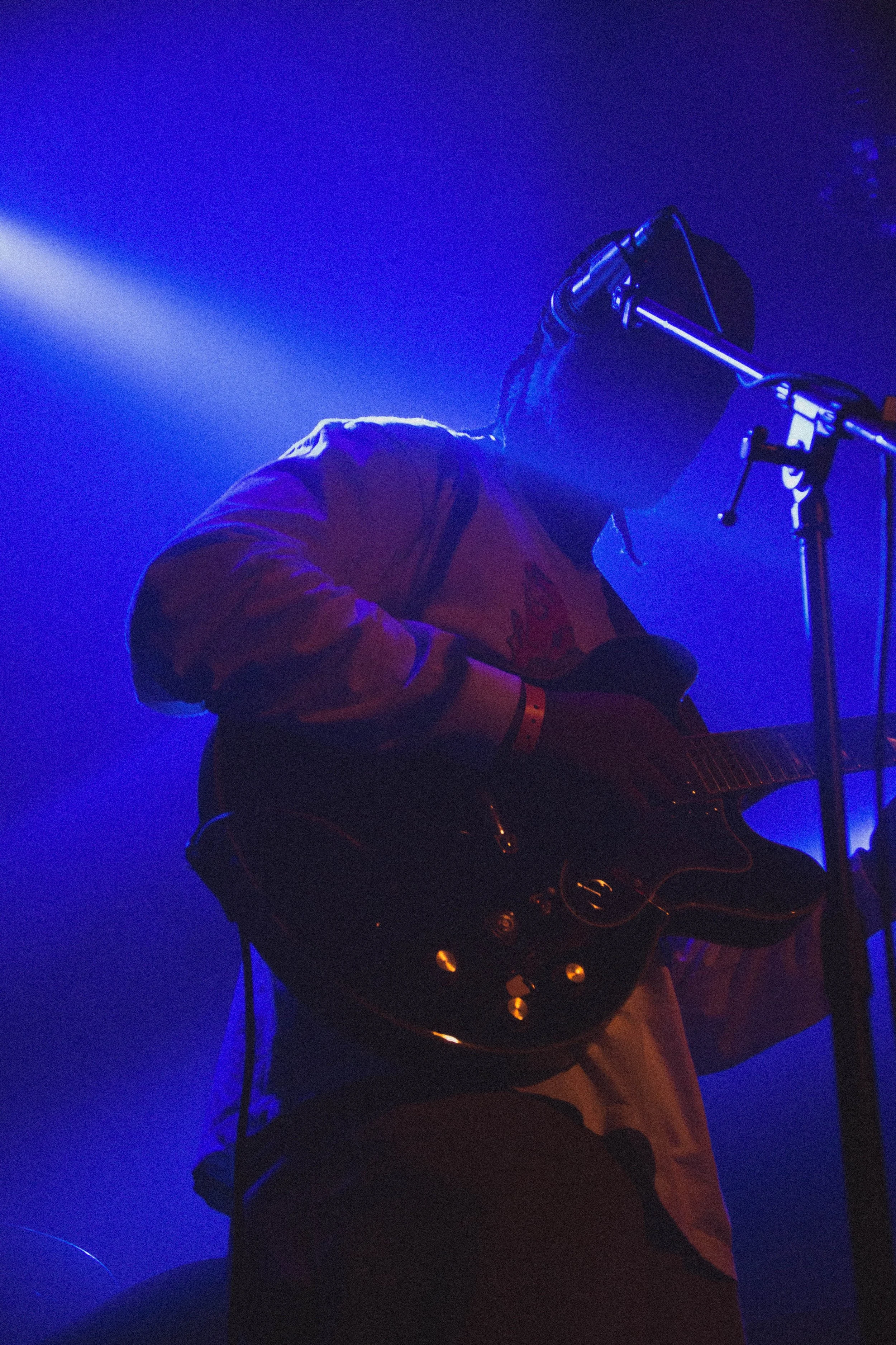 Musicien jouant de la guitare électrique sur scène avec un éclairage bleu et violet, portant un casque de musique.