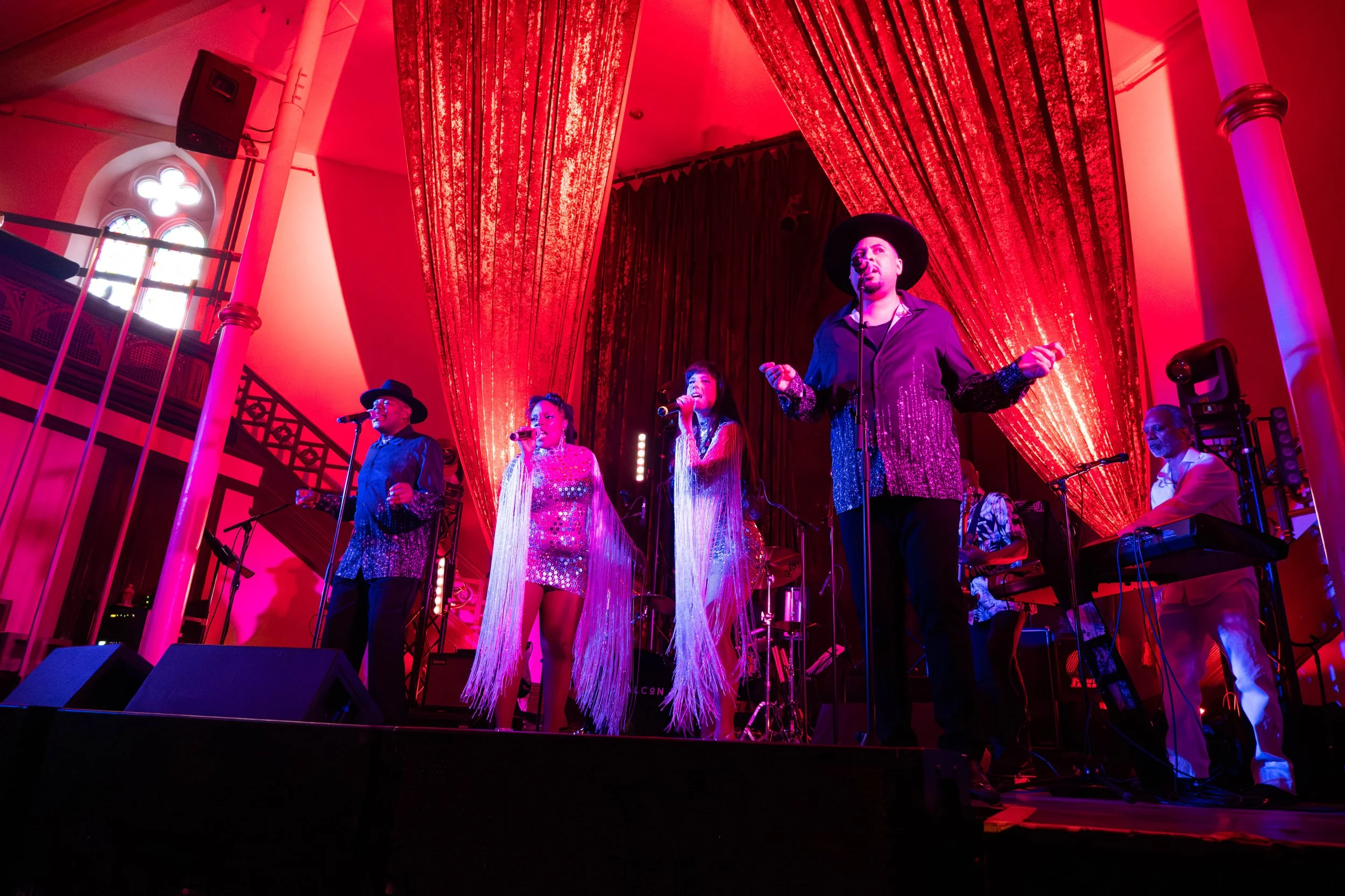 Un groupe de musiciens et chanteurs sur scène, sous un éclairage rouge, dans une salle avec des draperies rouges sur le fond.