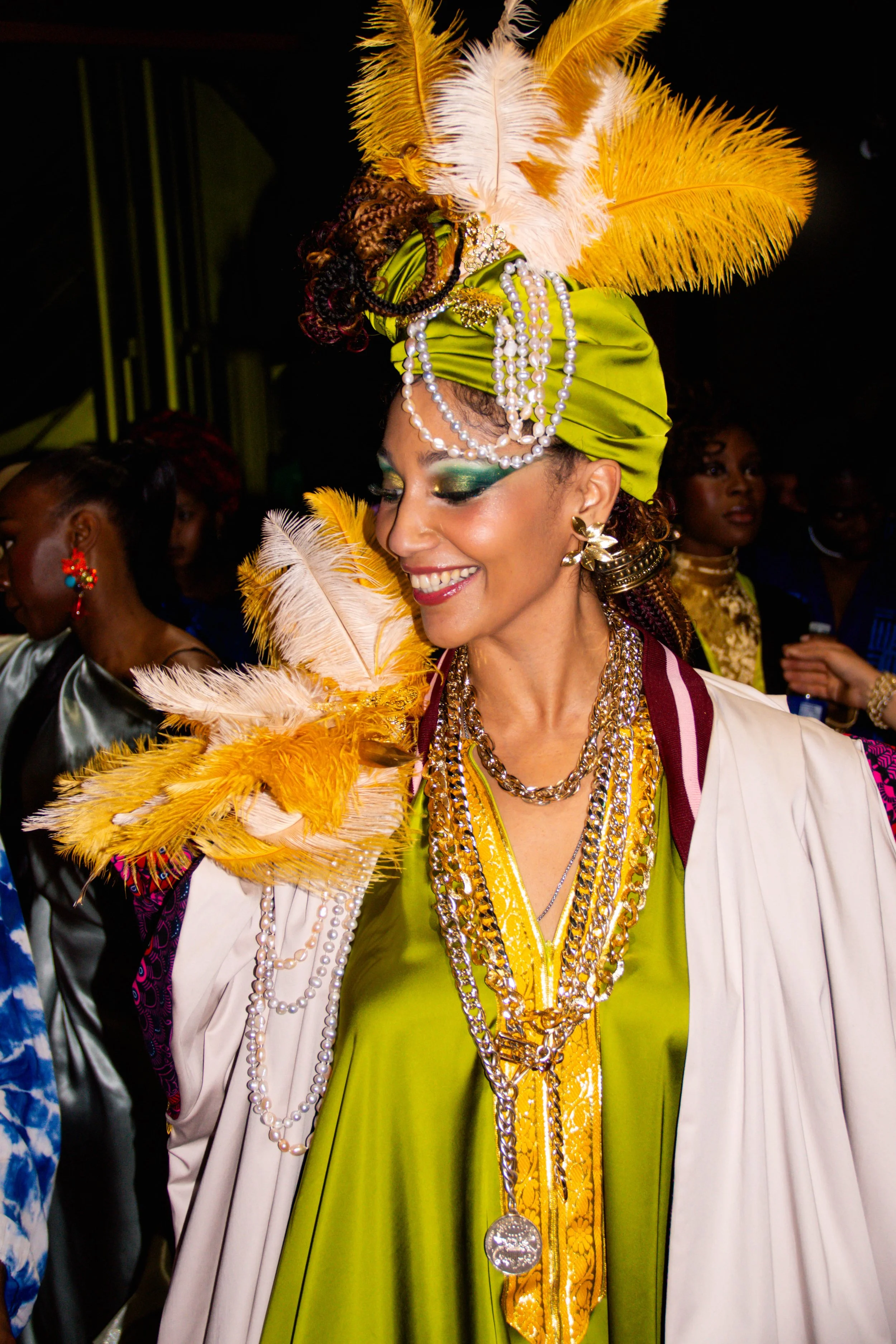 Une femme portant un chapeau orné de plumes jaunes et blanches, parée de bijoux en perles et de vêtements colorés, sourit lors d'une célébration ou d'un événement festif