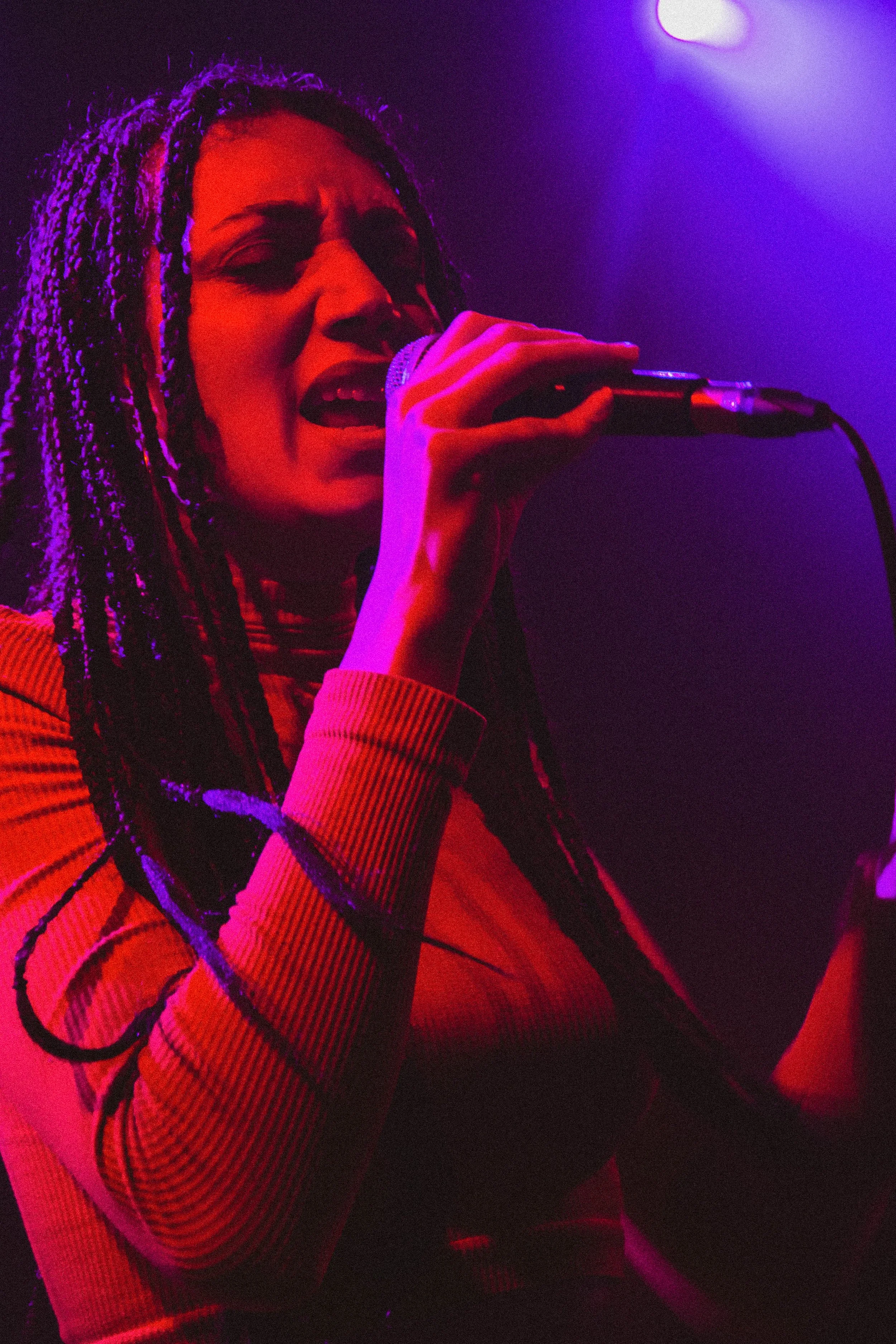 Une femme chante dans un micro sous des lumières violettes et rouges, ambiance de scène de concert.