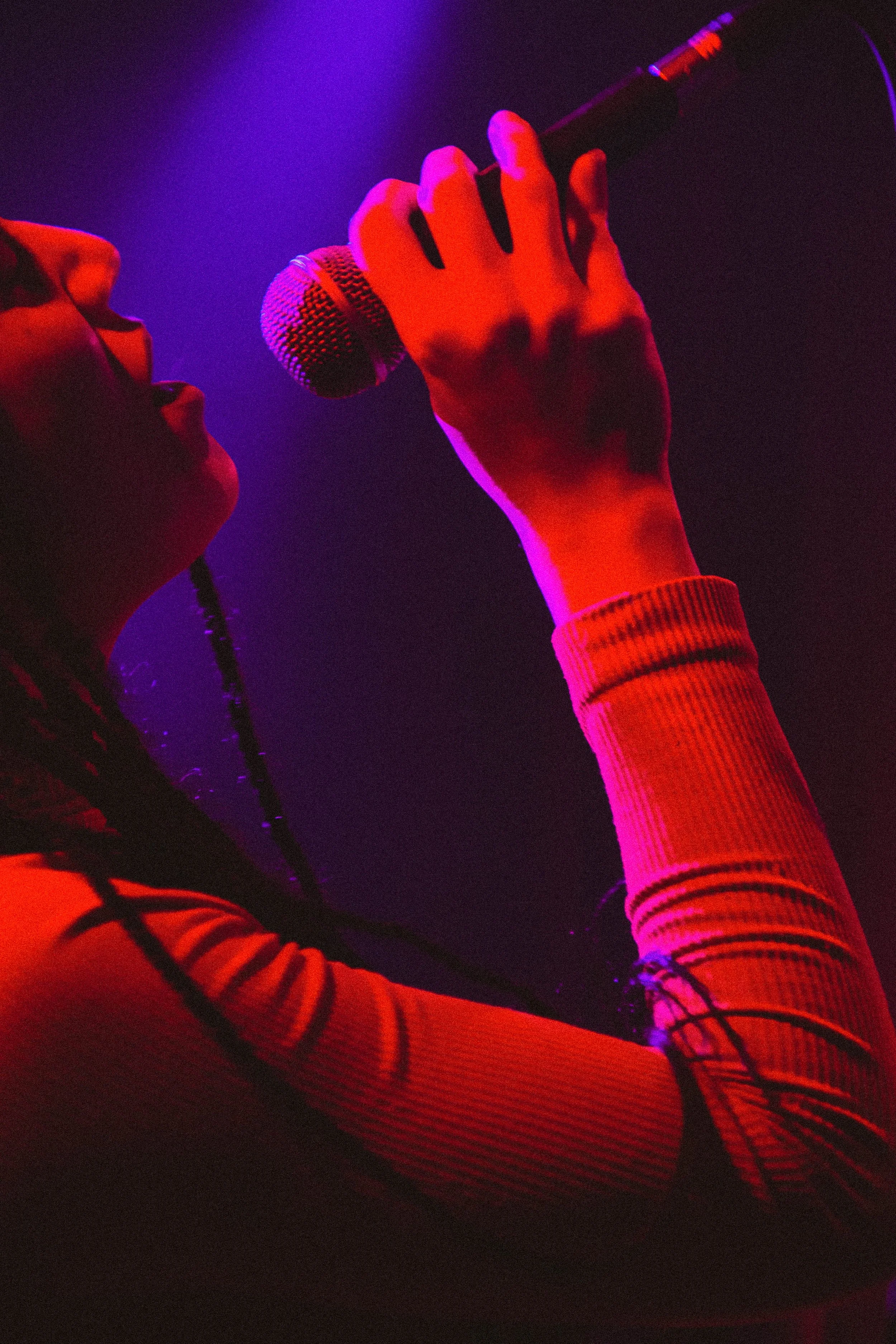 Une personne chante dans un micro sous un éclairage violet et rouge, portant un vêtement à rayures.