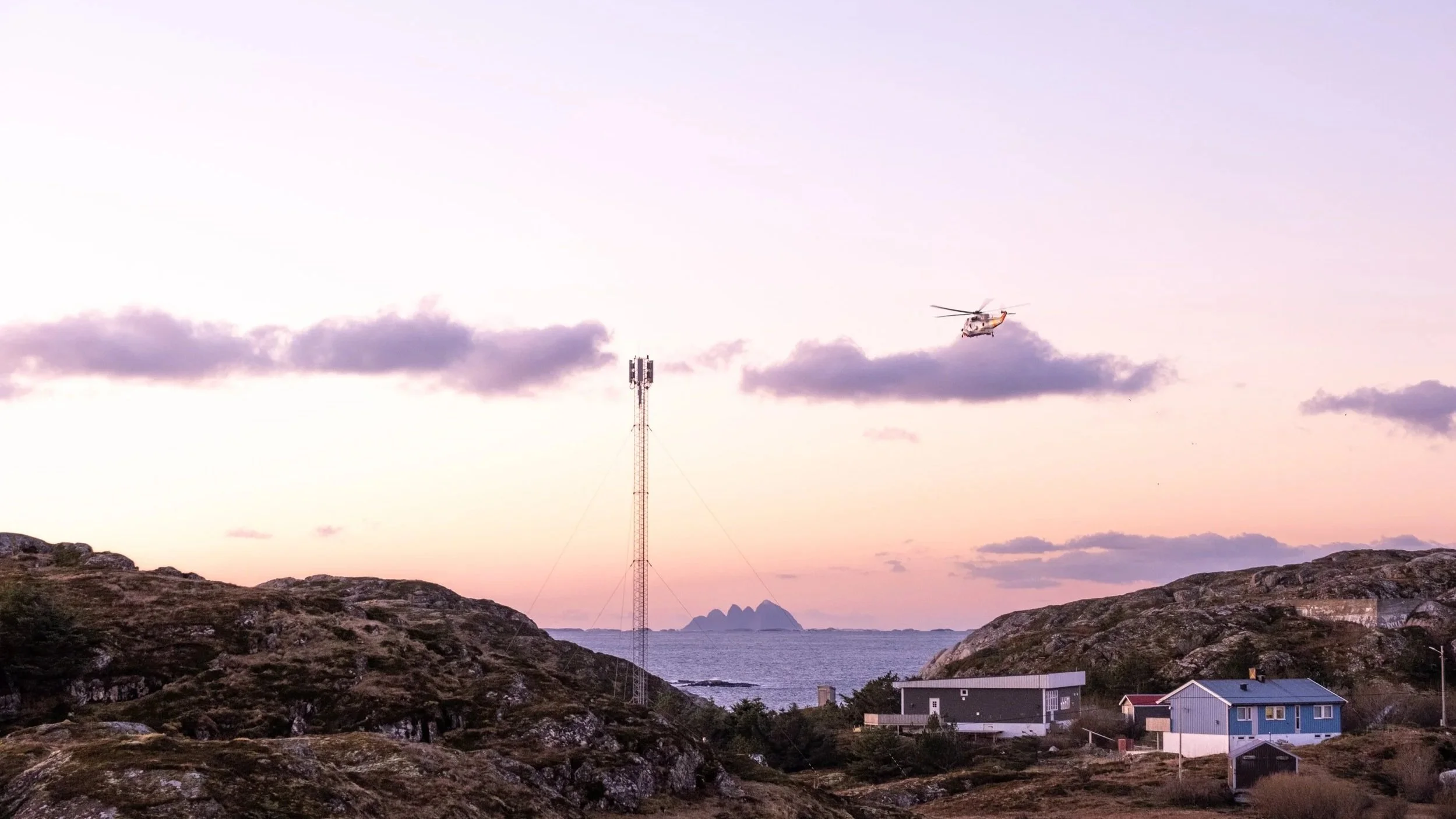 Scenic landscape at dusk with rocky hills, a few houses, a water tank, and a helicopter flying in the sky over the ocean and distant islands.