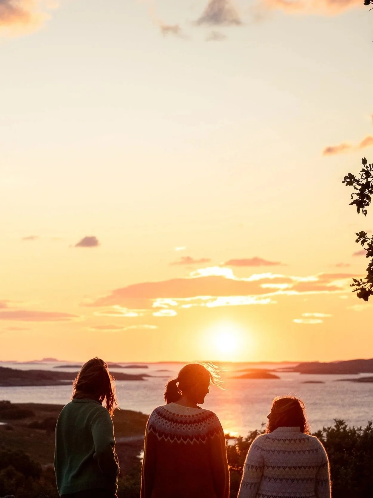 Three women standing outdoors during sunset, overlooking a body of water with small islands, wearing sweaters and enjoying the view.