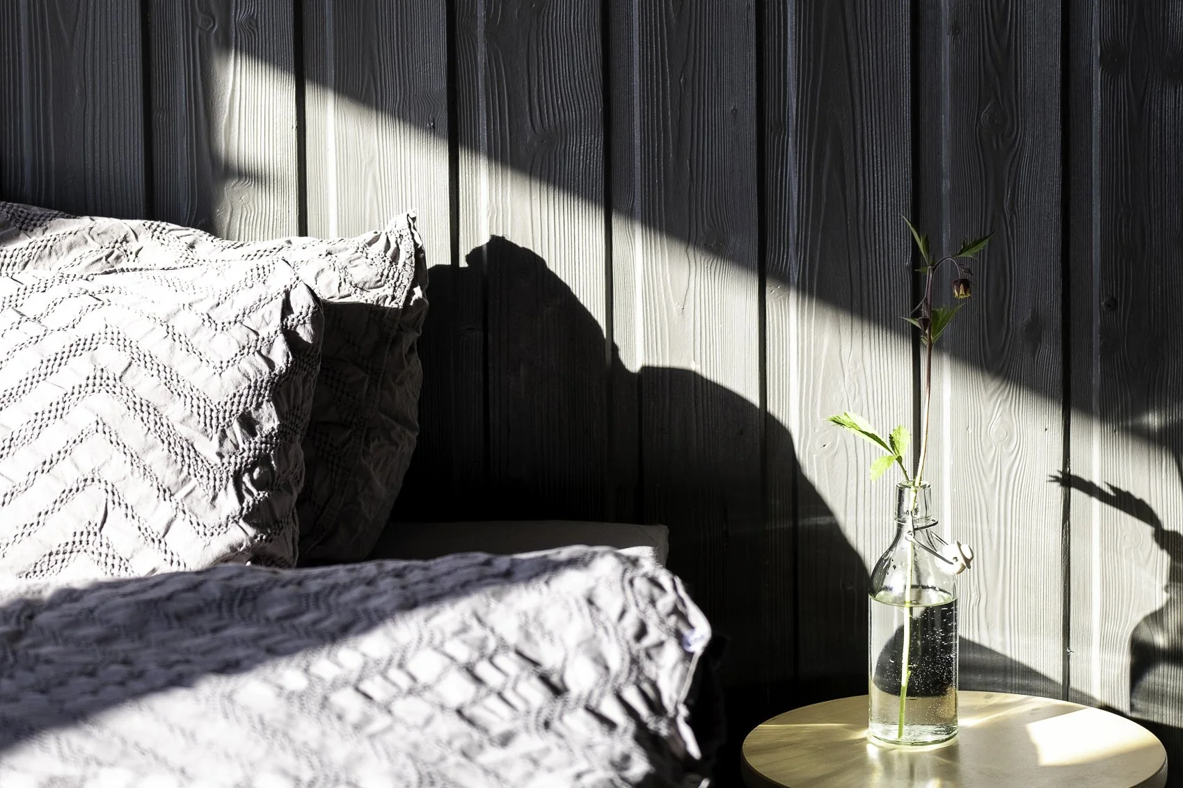 Sunlight casting shadows on a bed with textured gray bedding and pillows, a black wooden wall in the background, and a small round wooden table holding a clear glass vase with a single green plant stem and a dark purple flower.