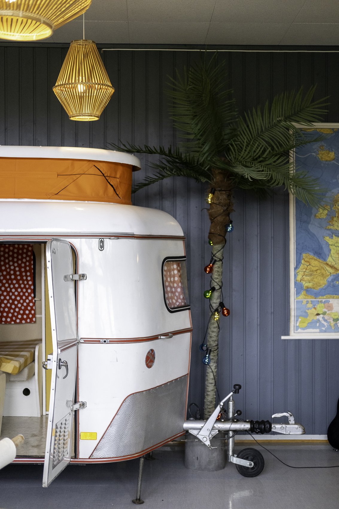 Inside a retro-style camper with a small door open, decorated with red and white polka dot curtains, a yellow and white striped bed. There is a large faux palm tree with string lights wrapped around the trunk, and a map on the wall behind it. The ceiling has wooden hanging lights.