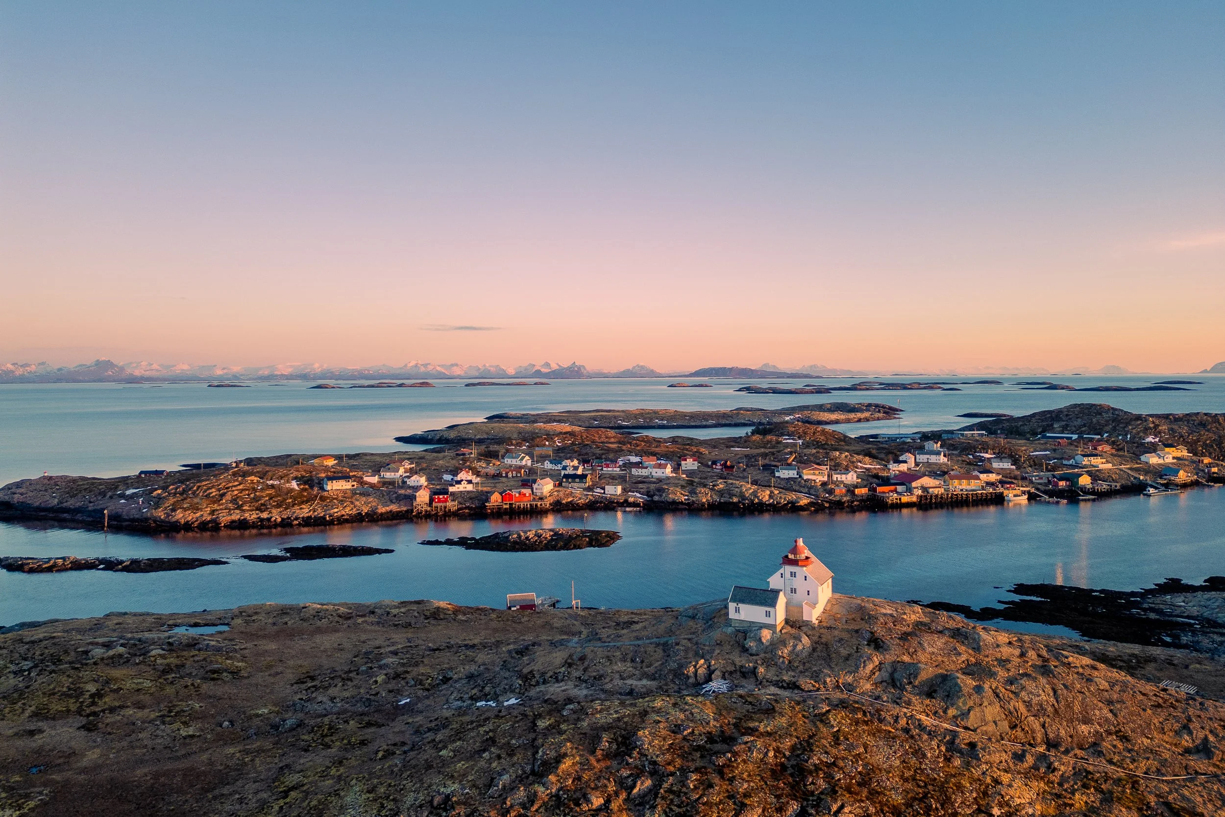 Scenic view of a coastal village with a white lighthouse in the foreground, surrounded by rocky terrain and water, under a pastel-colored sky at sunset.