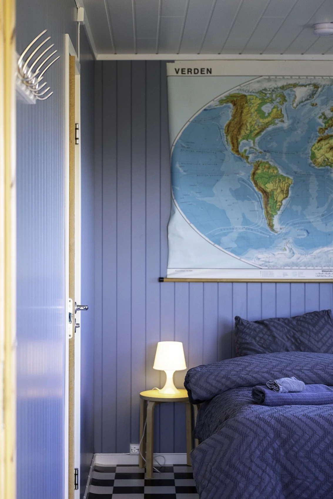A bedroom with blue-paneled walls, a bed with dark bedding, a small table with a white lamp, a world map on the wall, and a partially open door.