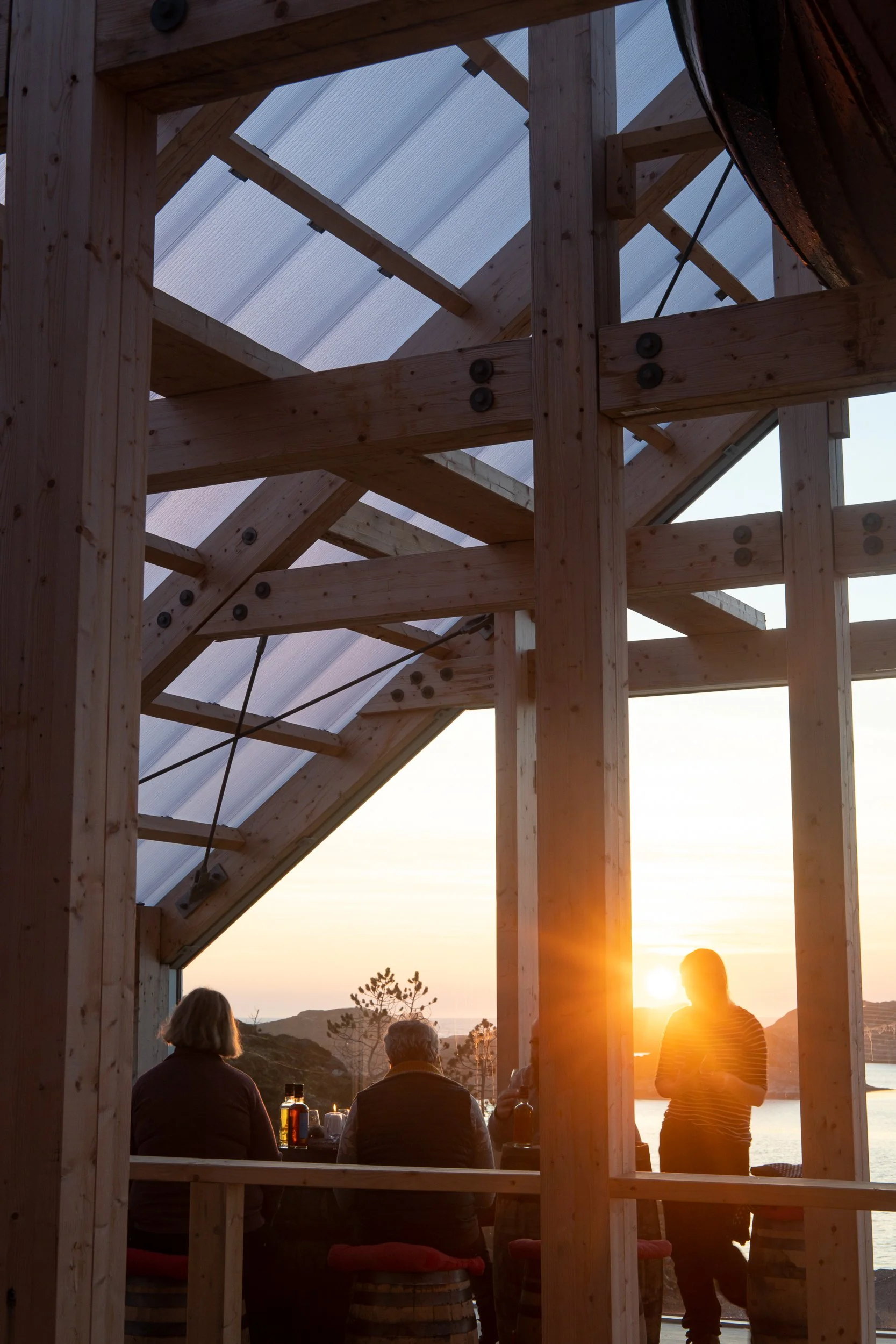 People sitting at a table inside a partially constructed wooden building with a view of the sunset over a body of water.