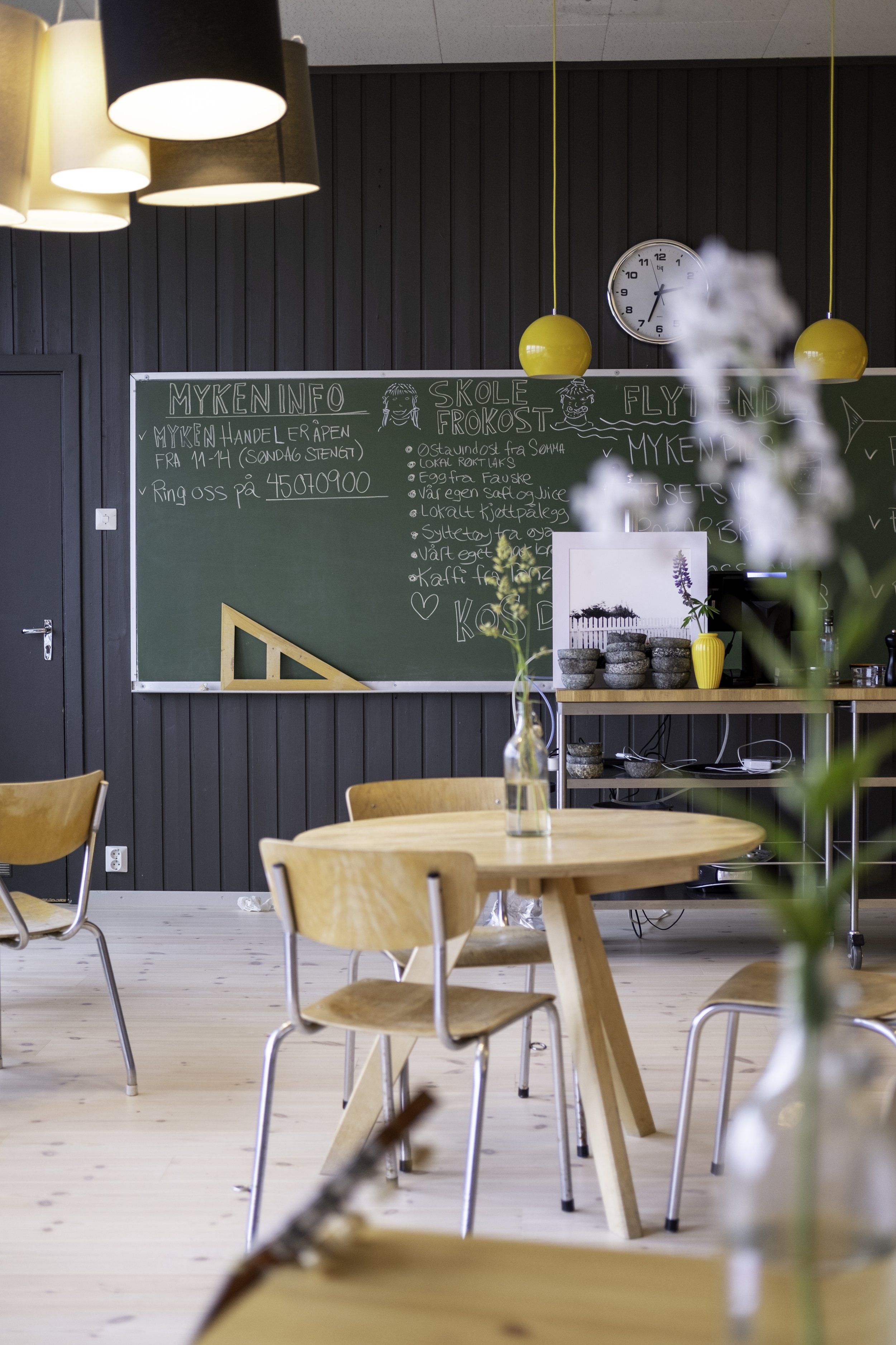 Interior of a cozy cafe with wooden tables and chairs, a chalkboard menu on a dark wall, pendant lights, and decorative vases with flowers.