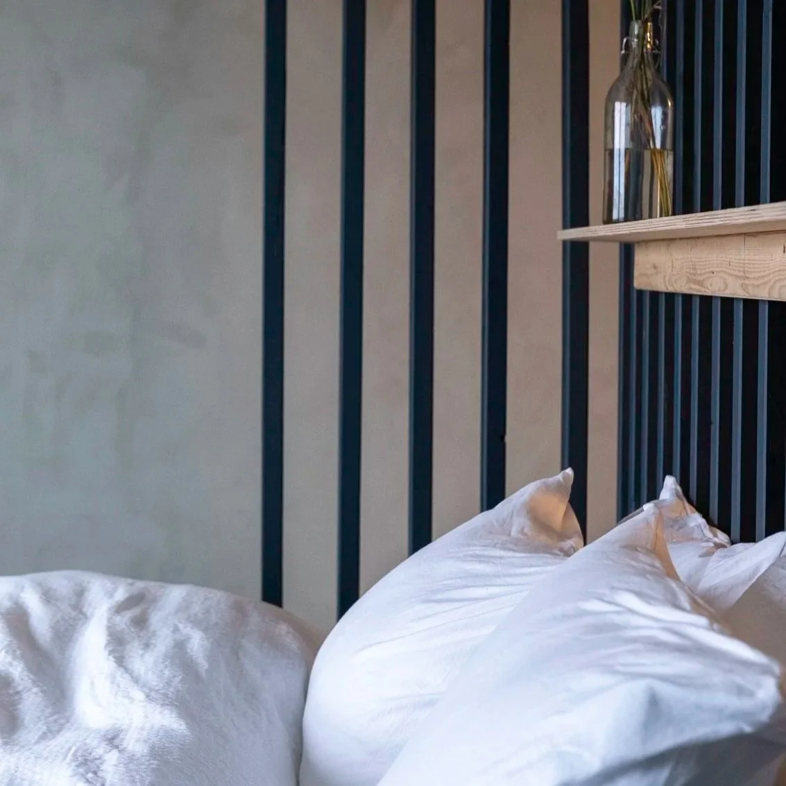 Close-up of a bed with white pillows and bedding, against a wall with black vertical metal bars and a wooden shelf with a glass vase containing yellow flowers.