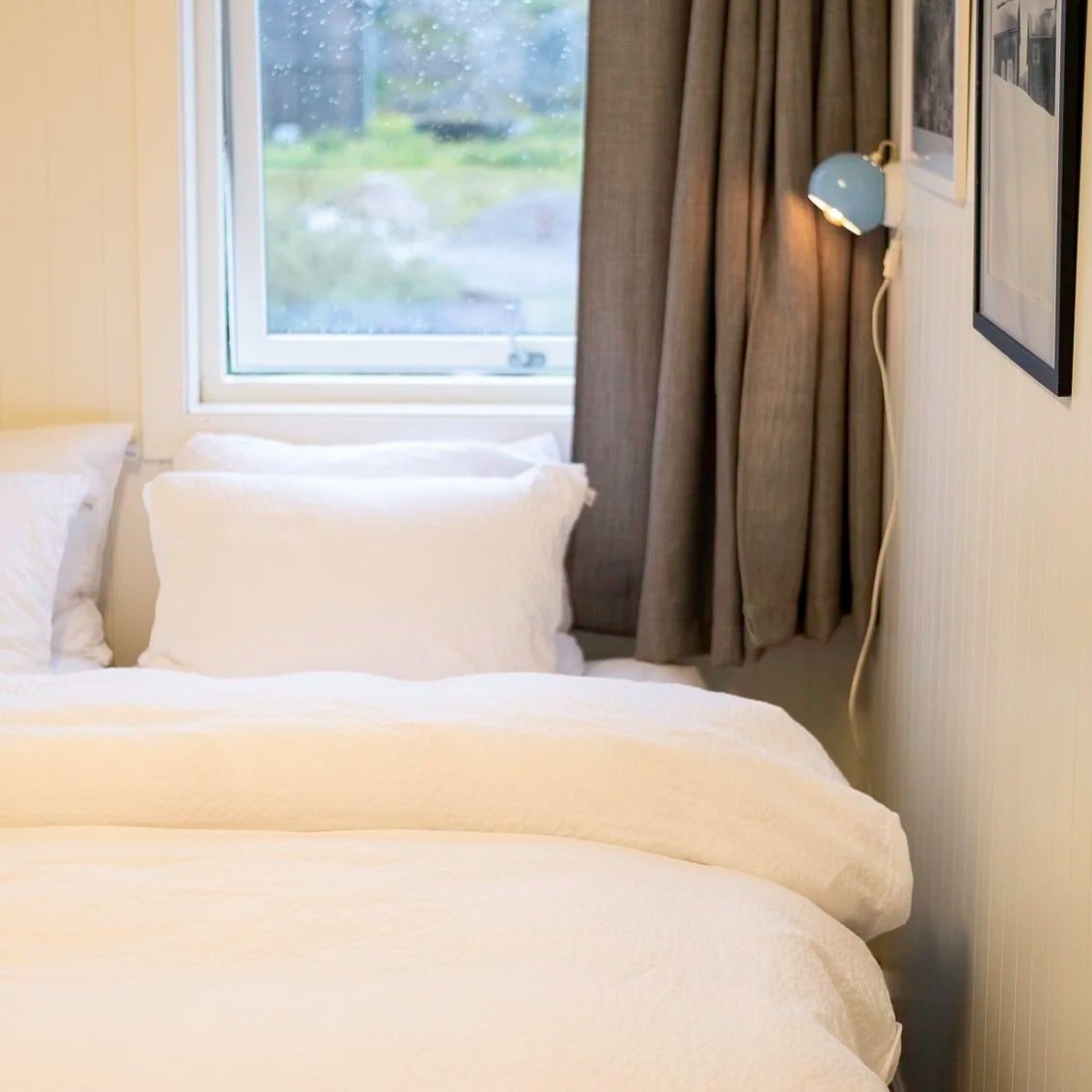 A cozy bedroom with two white beds, a window with partly open curtains showing a green outdoor view, and a wall-mounted lamp next to framed artwork.